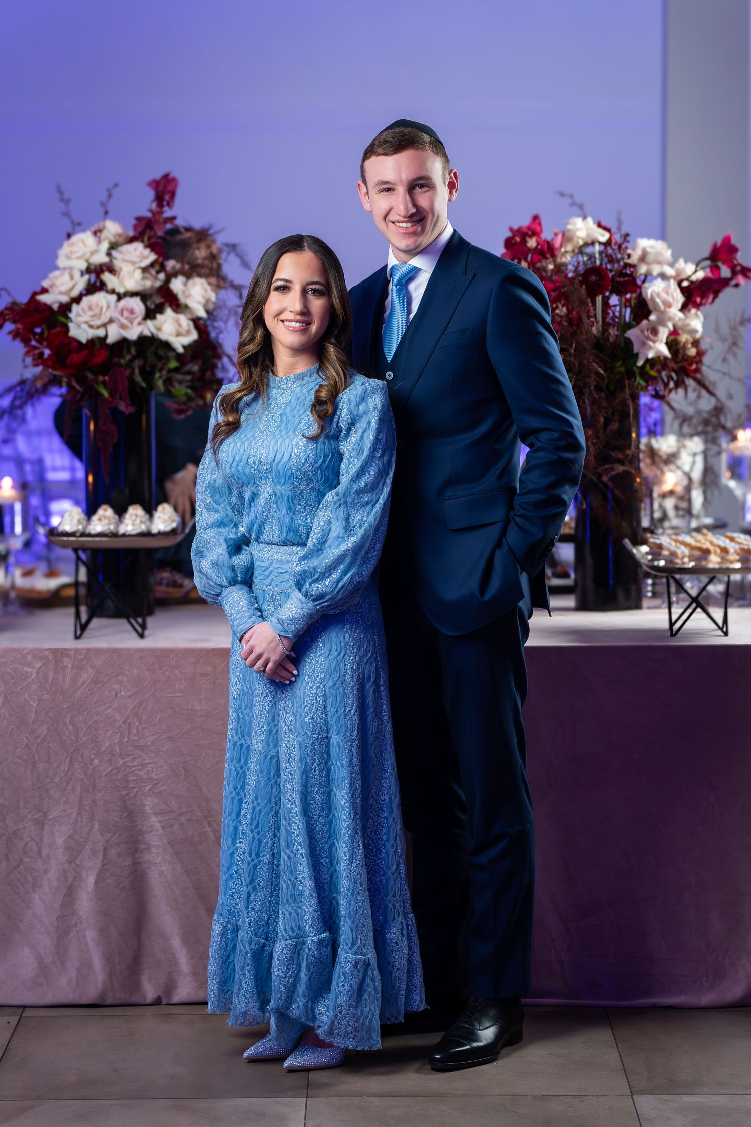 A couple poses at an elegant event with floral arrangements and dessert table in the background.