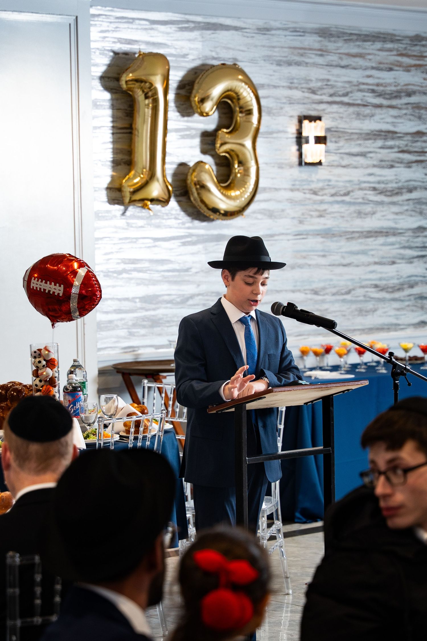 A boy in a suit and hat speaks at a podium during his Bar Mitzvah celebration with gold number 13 balloons.