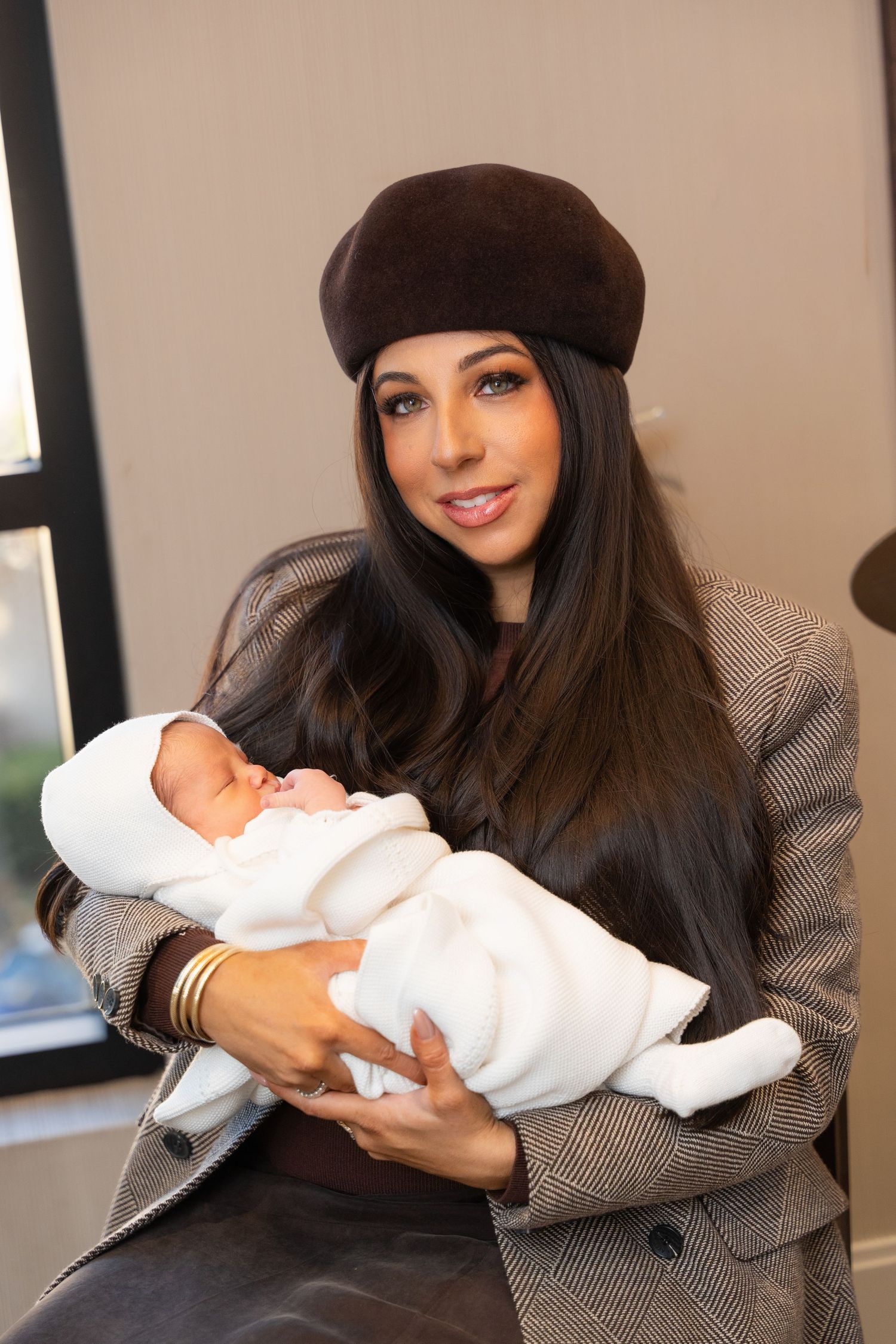 A woman in a gray blazer and black beret holds and gazes lovingly at a newborn baby wrapped in white blankets.