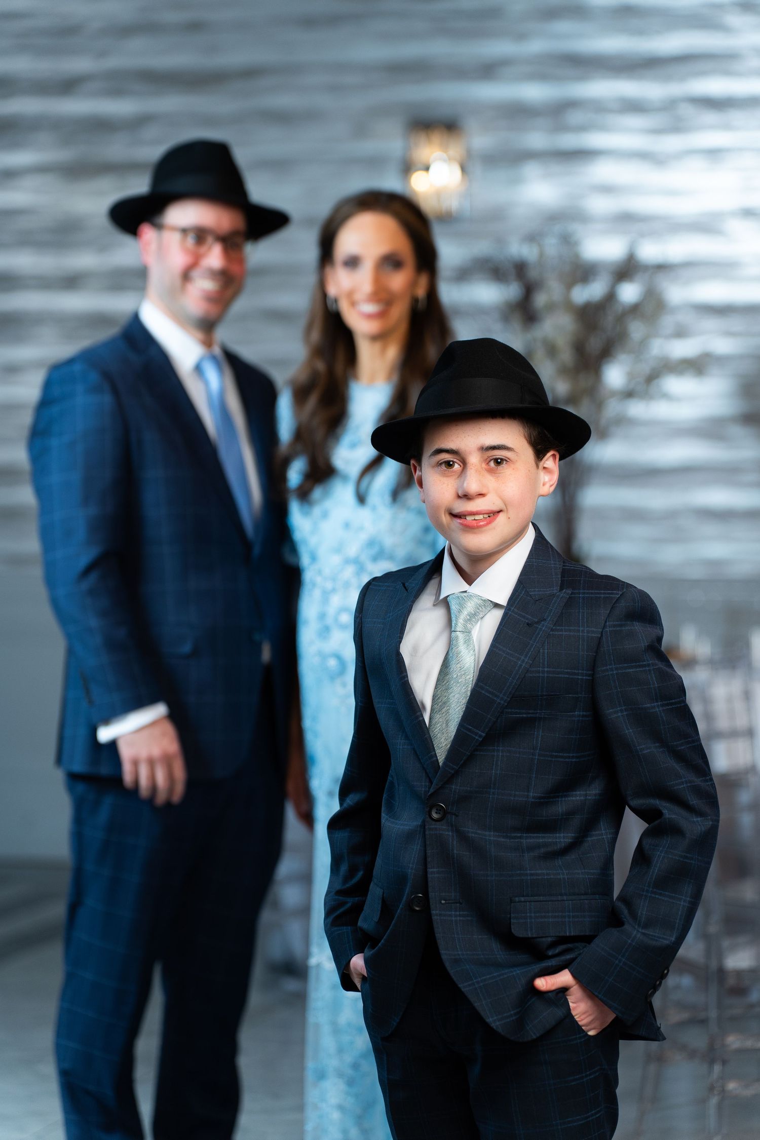 Boy in suit and hat poses with parents at formal Jewish Bar Mitzvah celebration event.