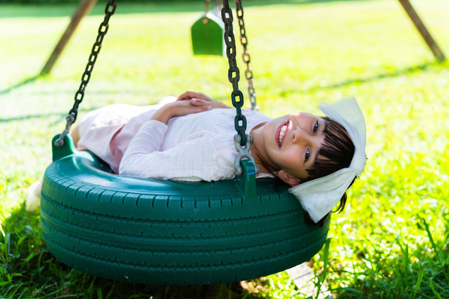 Young girl smiling while lying in a green tire swing outdoors on a sunny day.