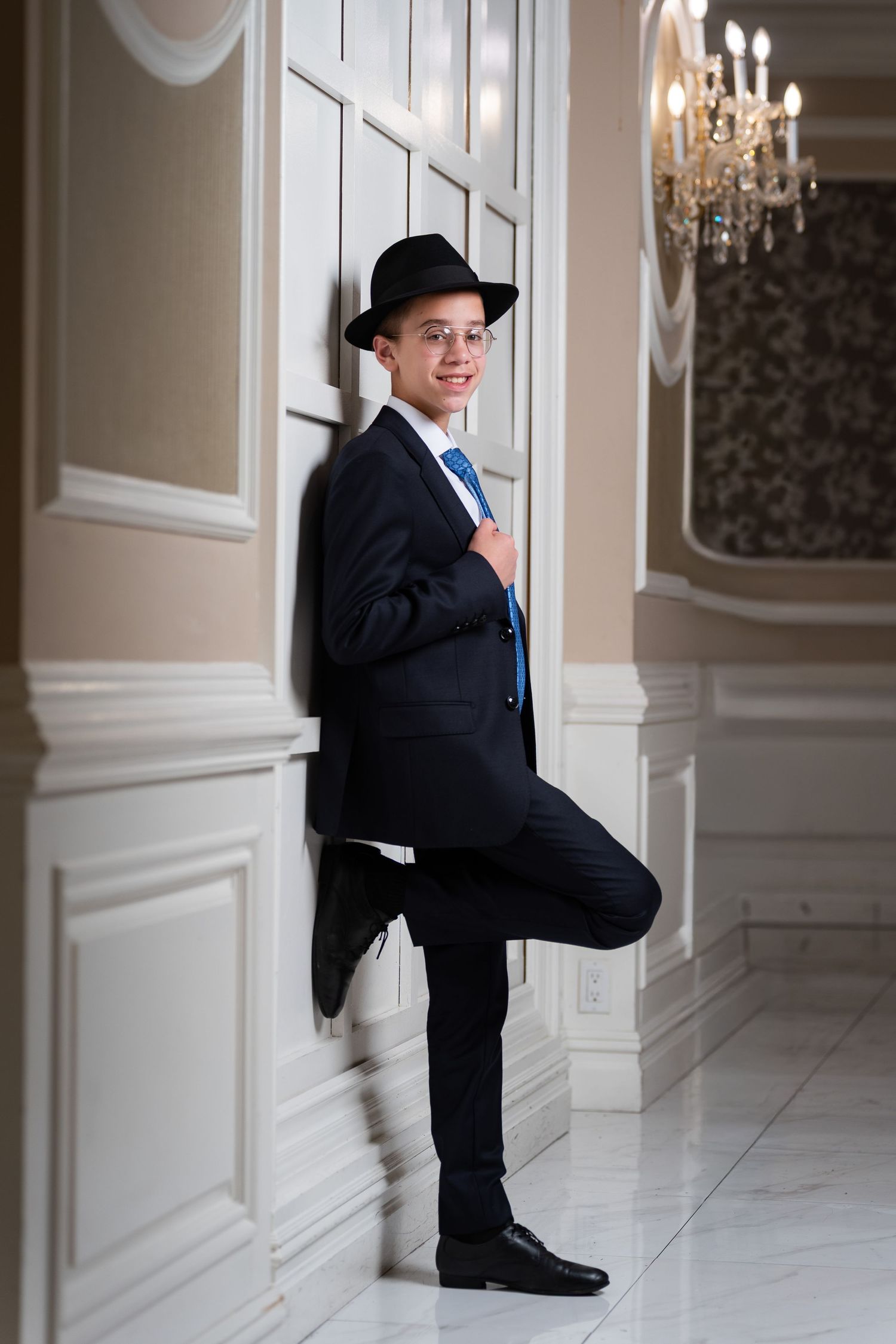 Young man in black suit, hat, and glasses leans against white wall in elegant room with chandelier.