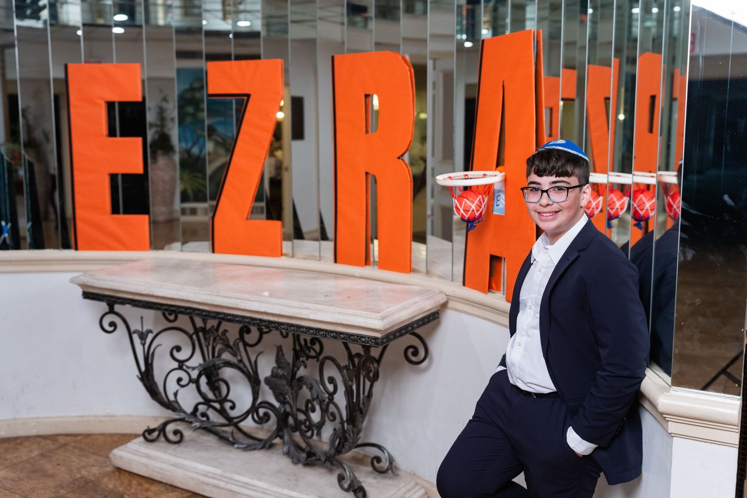 Young man in suit poses beside large orange EZRA letters at a Bar Mitzvah celebration venue.