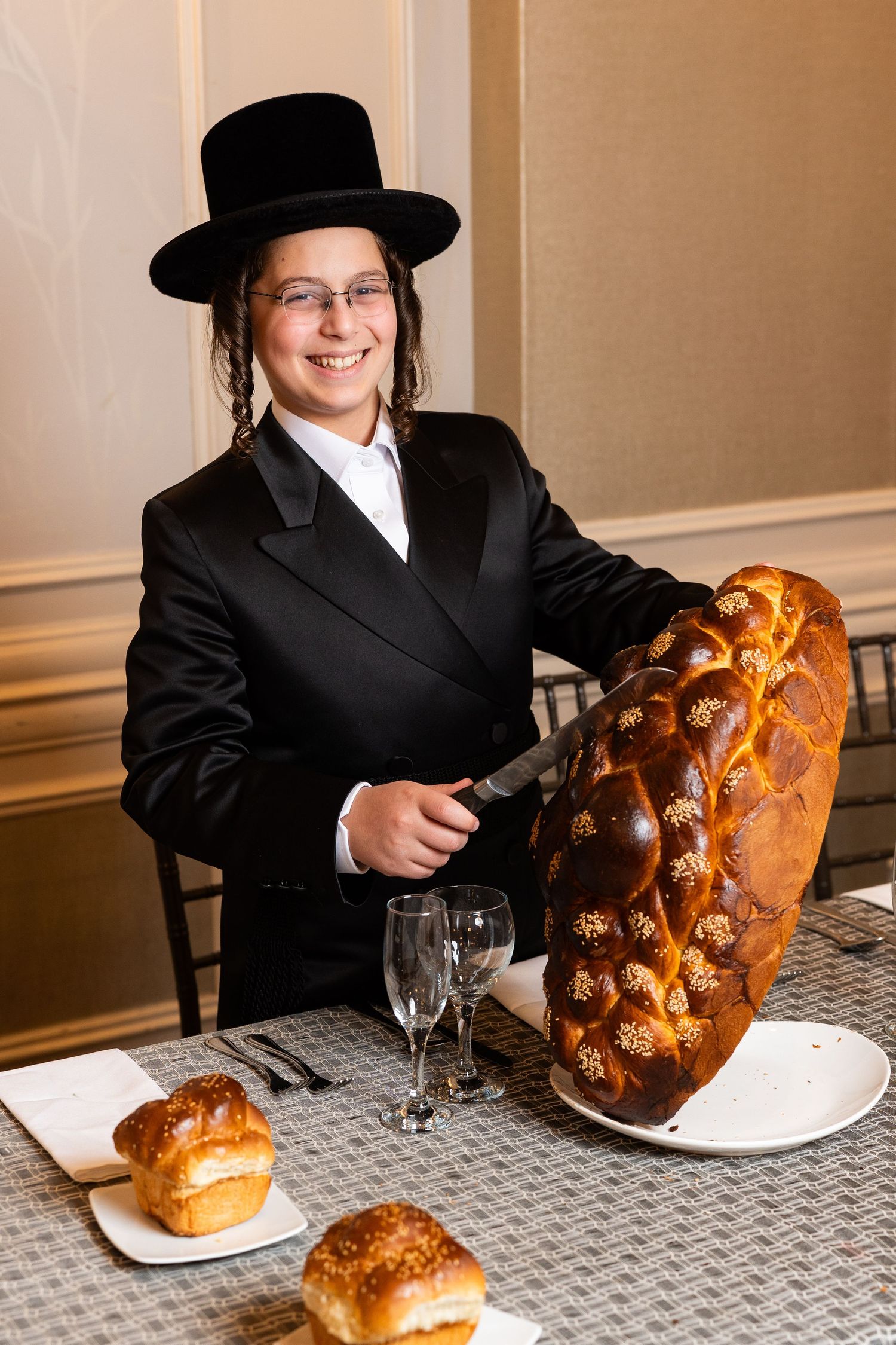 A person in traditional Jewish attire sits at a table with a freshly baked challah bread loaf.