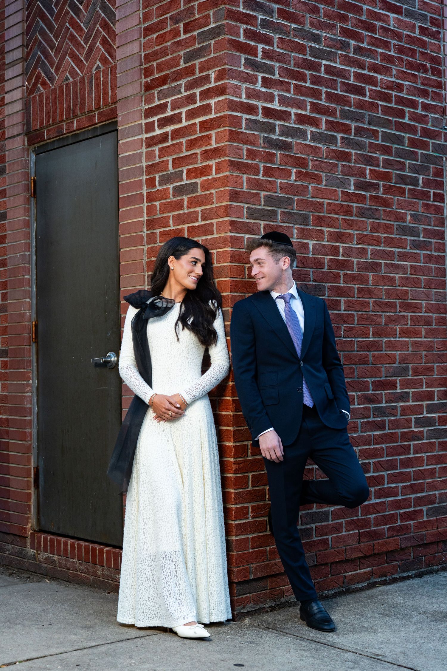 A couple poses playfully against a brick wall, with the woman in a white lace dress and the man in a dark suit.