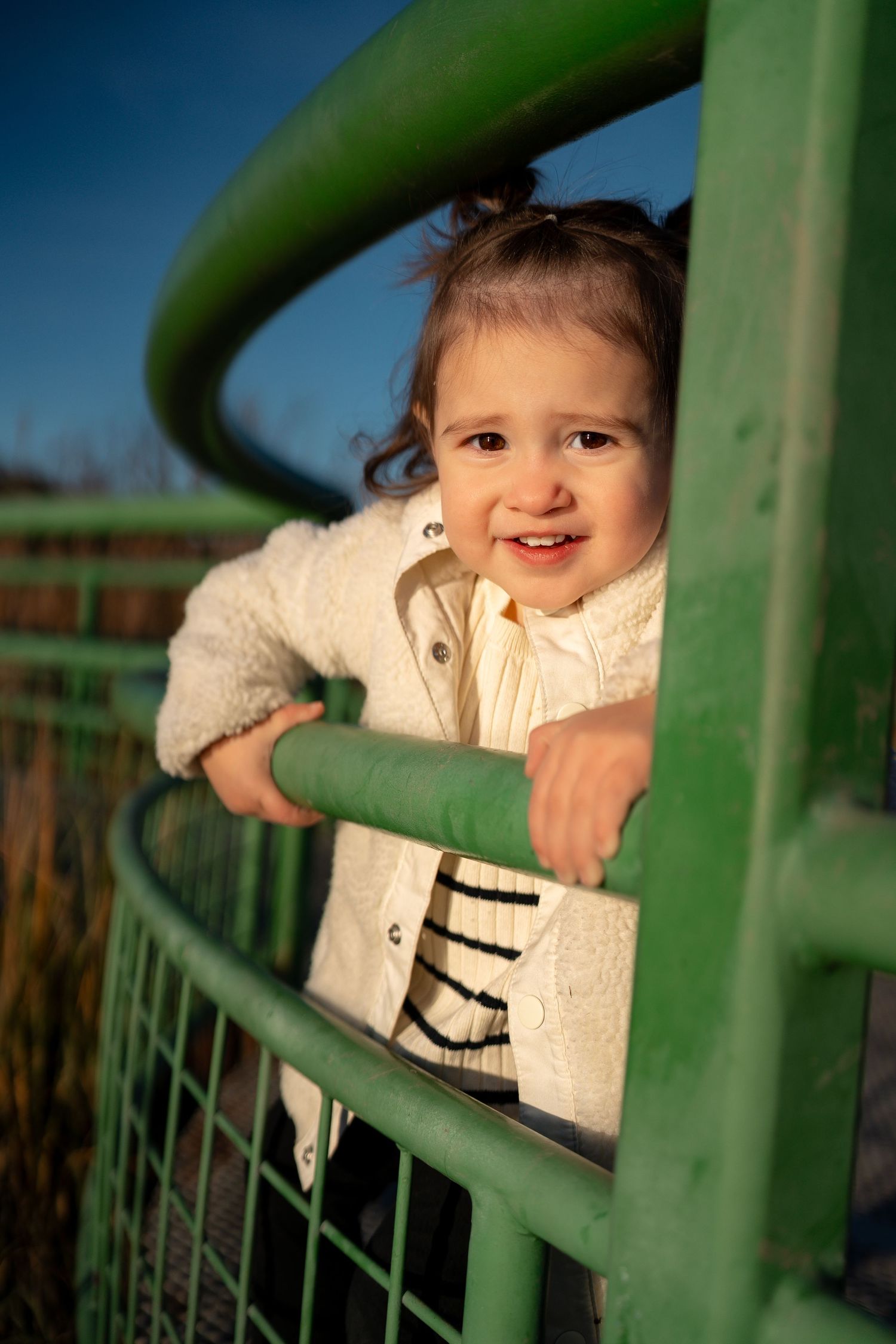 Child peeks playfully through green metal bars with a bright smile in outdoor setting.
