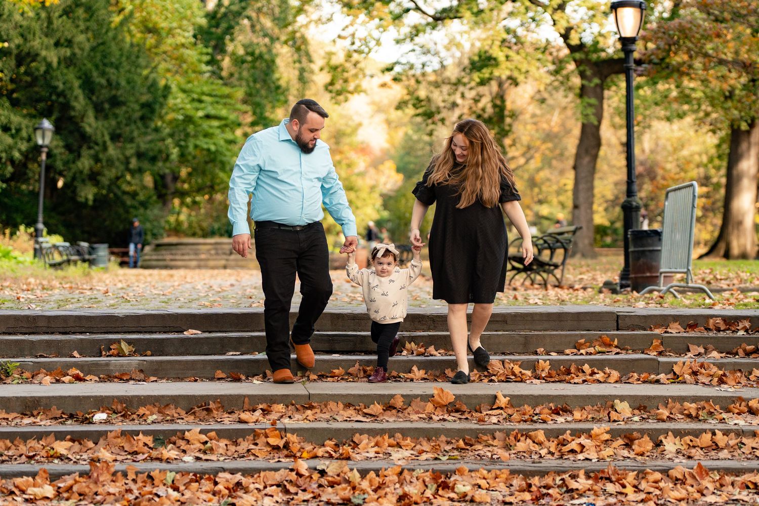 Couple walking a small dog on autumn leaf-covered steps in a park with colorful fall foliage.