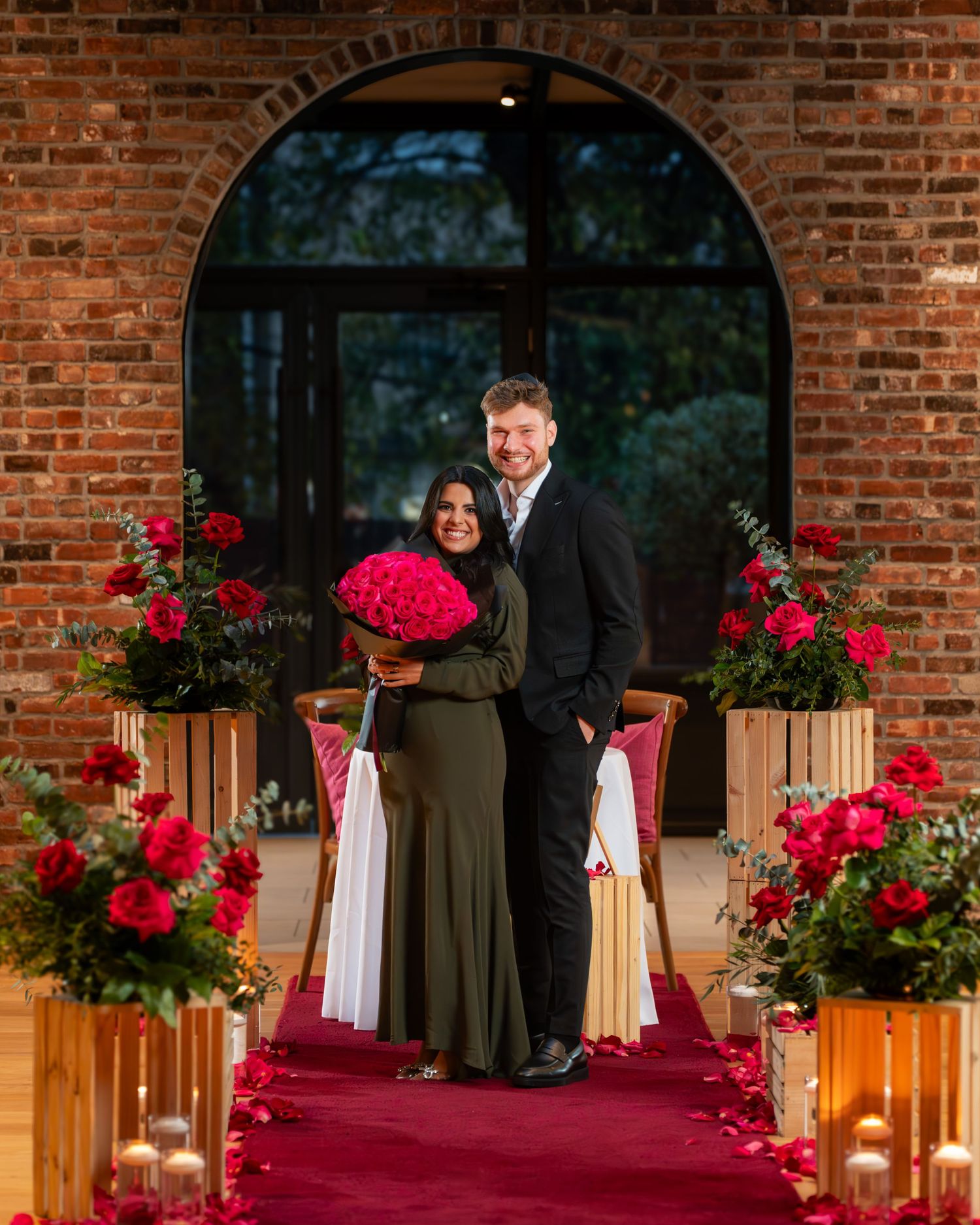 Elegant wedding ceremony setup with red roses and white fabric decorations against a rustic brick wall backdrop.