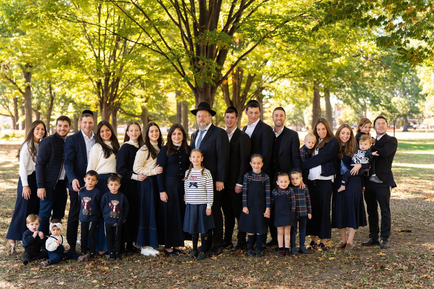 Large family group portrait outdoors in autumn park with yellow trees, dressed in formal navy and black attire.