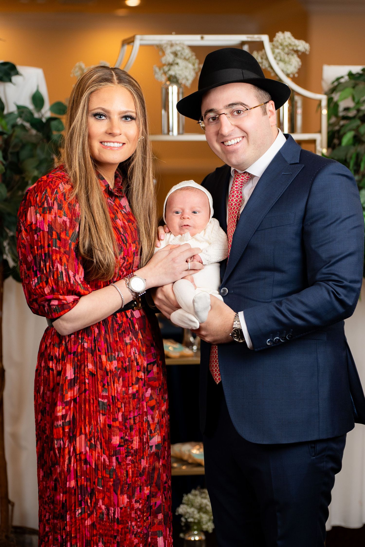 A couple in formal attire poses with a baby in a warmly lit indoor setting, the woman wearing a festive red dress.