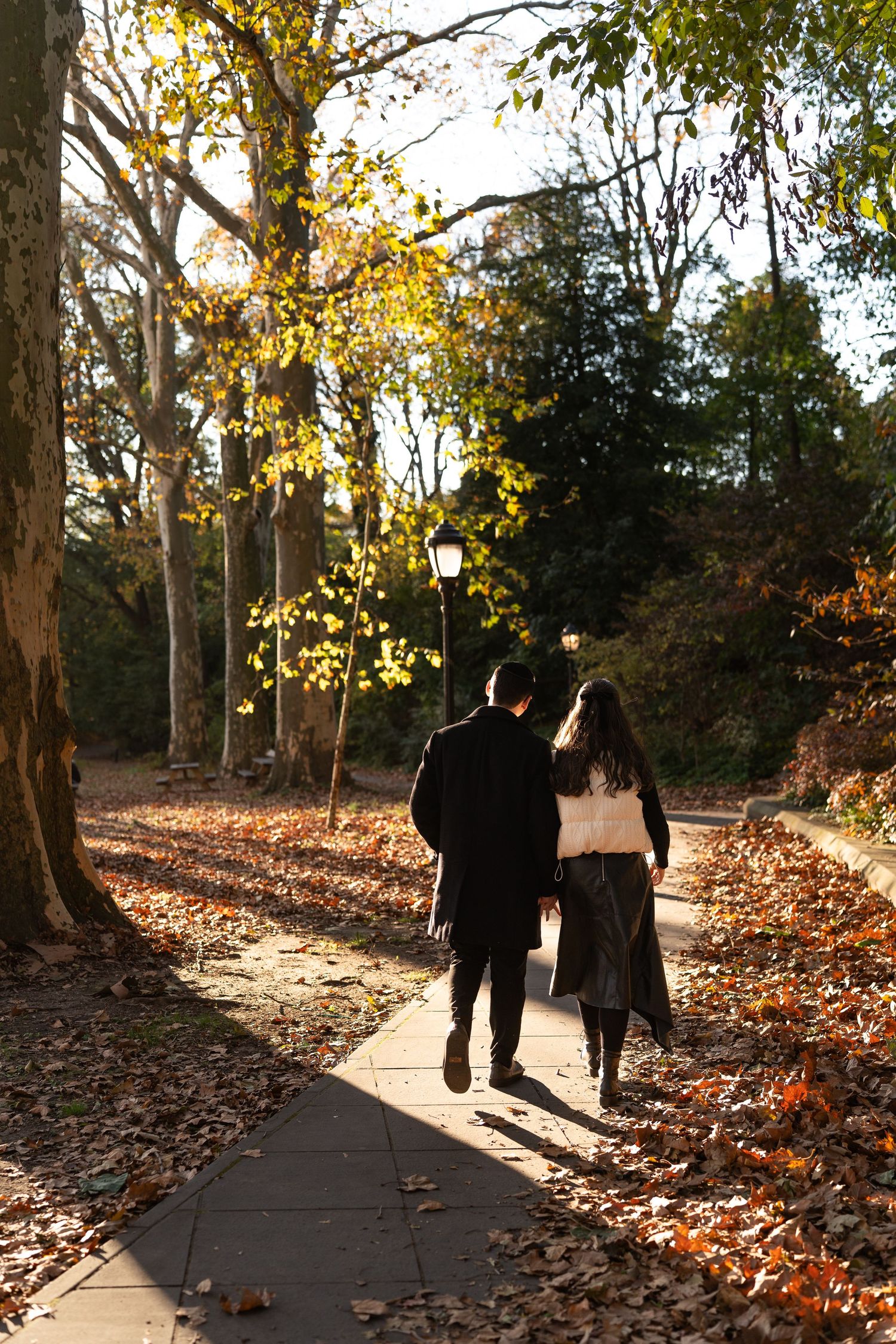 Couple walking hand in hand on a leaf-covered park path surrounded by autumn trees in golden sunlight.