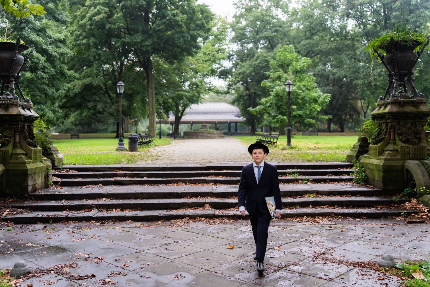 Person in suit and hat walking in a lush green park with stone steps and gazebo backdrop.