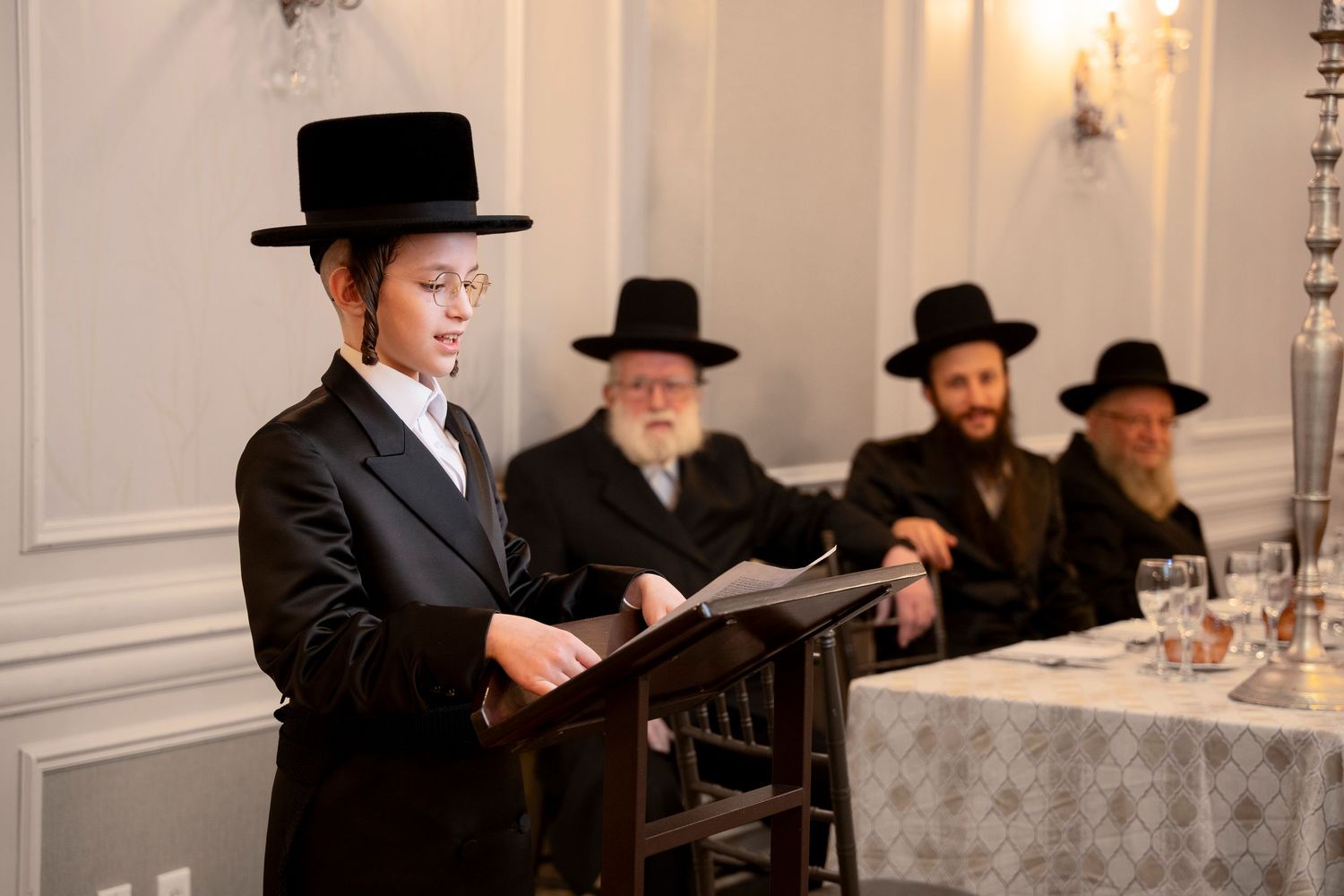 A group in traditional Orthodox Jewish attire reads from texts while seated at an elegant table with white tablecloth.