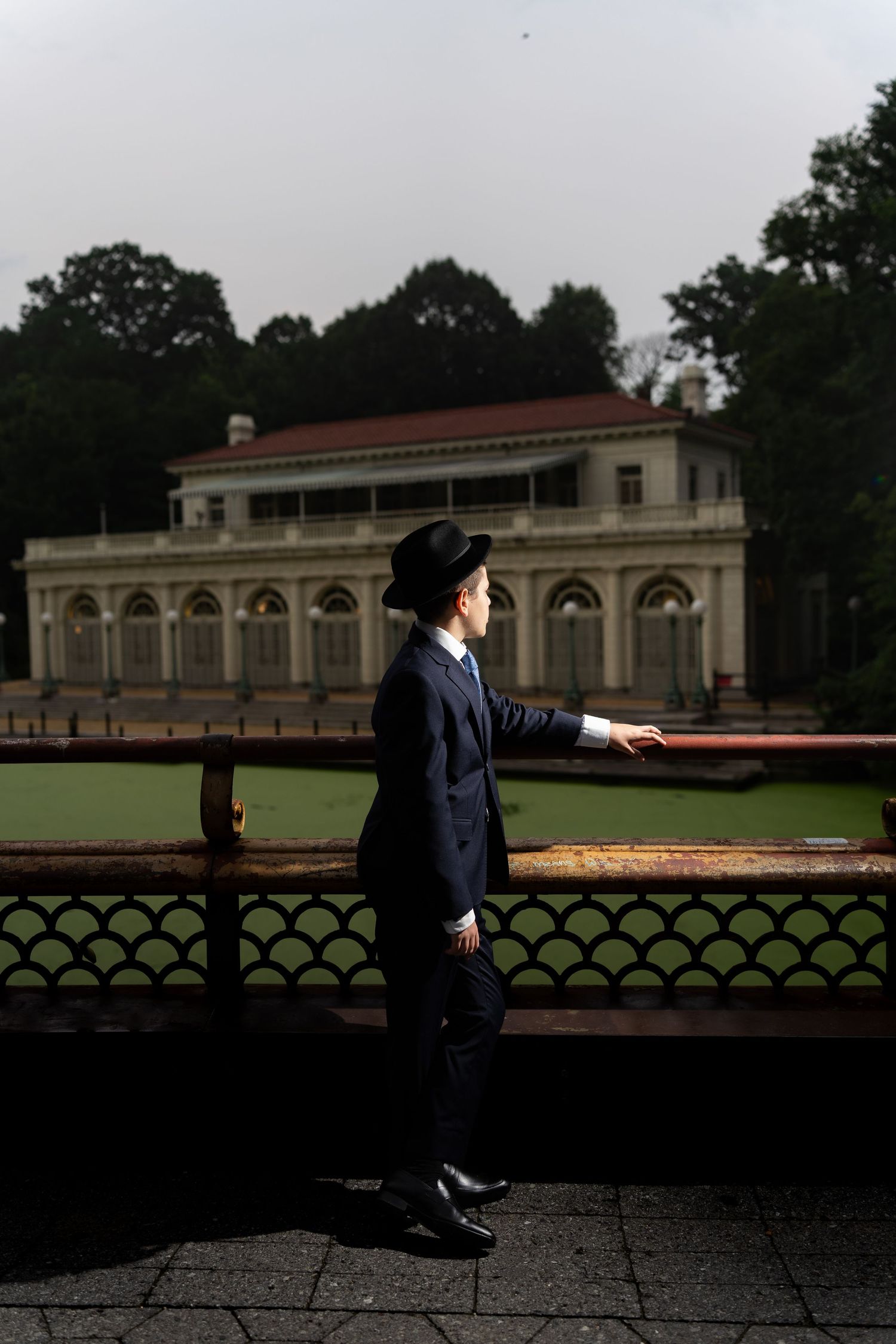 A man in a black suit and hat stands by an ornate fence near a green pond with a historic building behind.