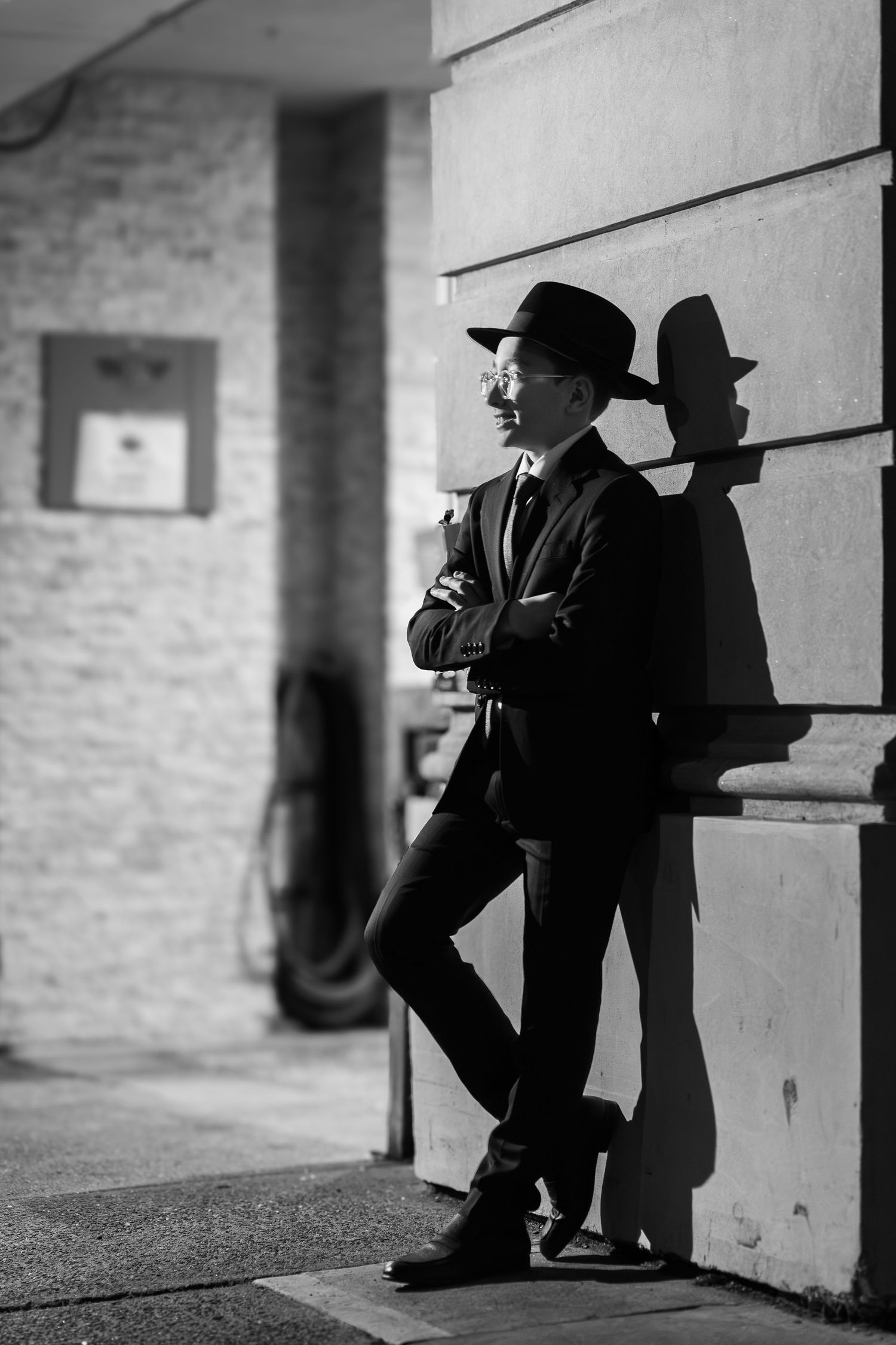 Black and white photo of a stylish man in a suit and hat leaning against a wall with arms crossed.