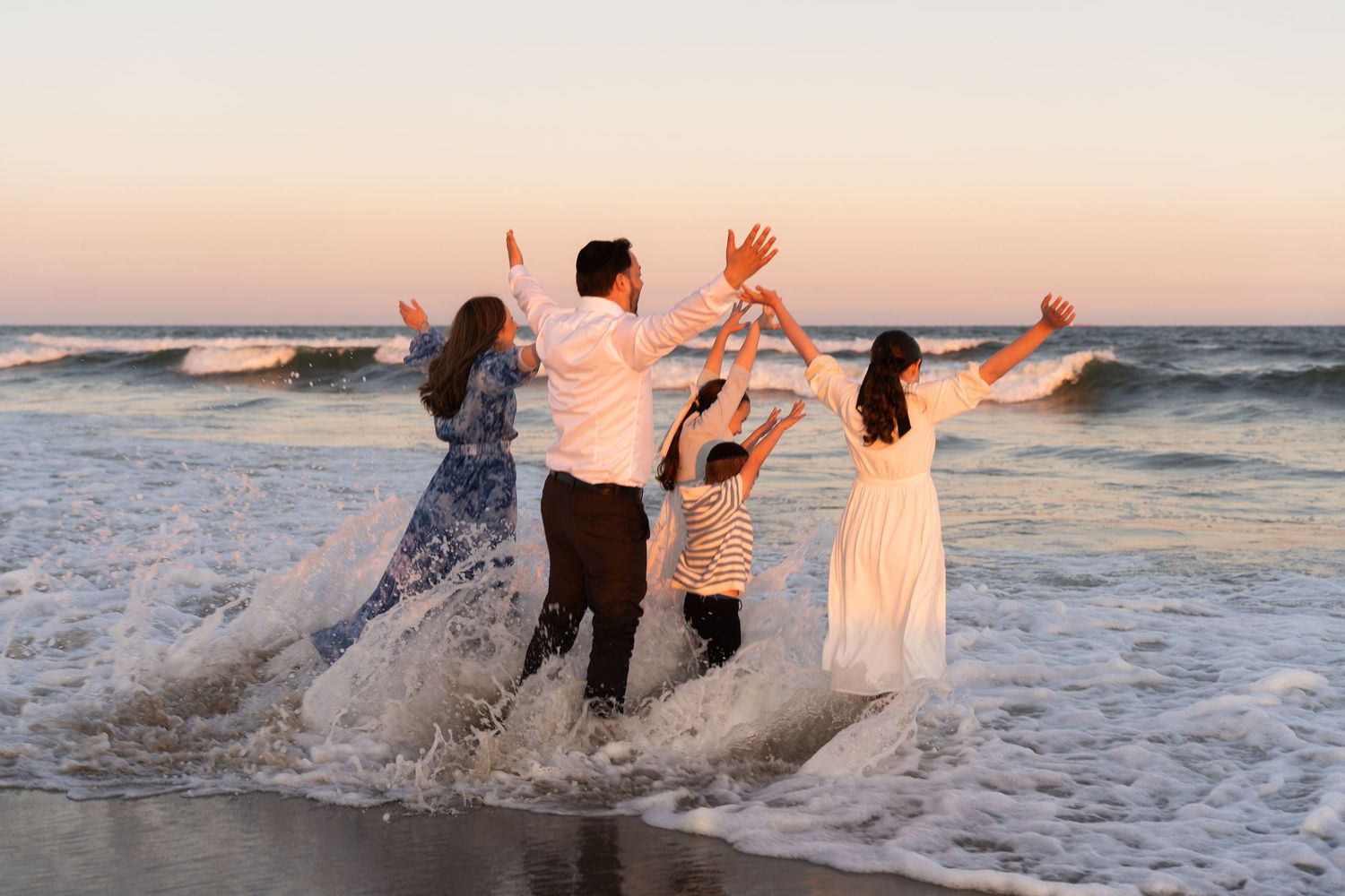 A group splashes through ocean waves at sunset wearing formal attire during a beach photo session.