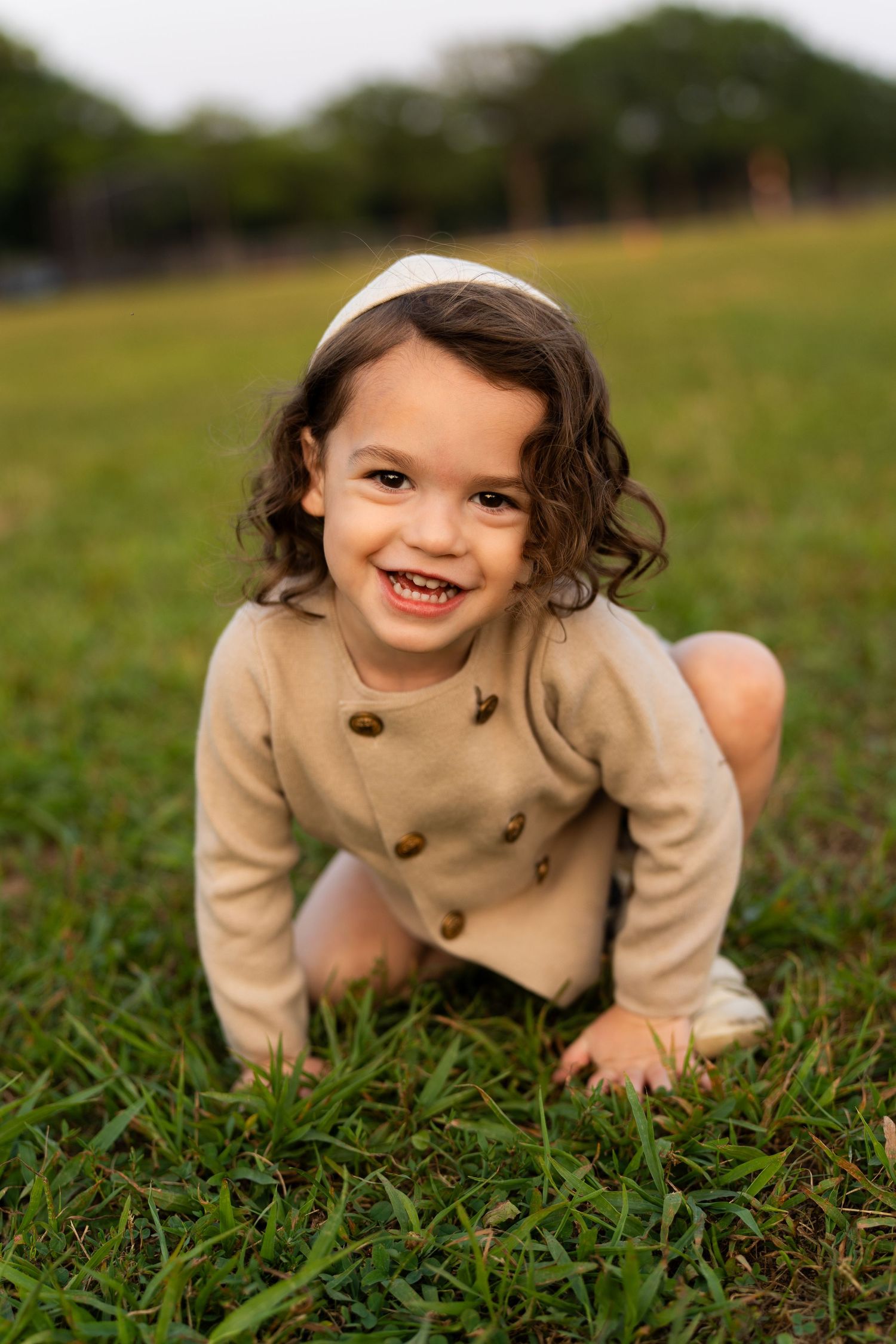 A child in a beige coat crawls and plays in the grass on a sunny day.