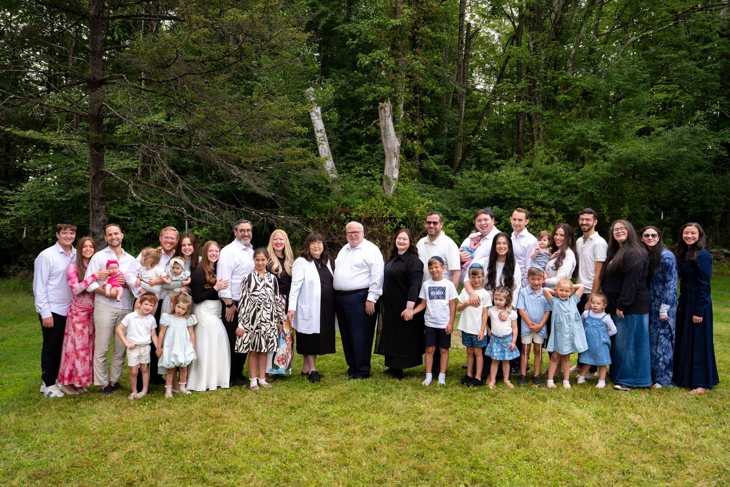 Large multigenerational family group portrait outdoors on green lawn surrounded by lush trees, dressed in formal attire.