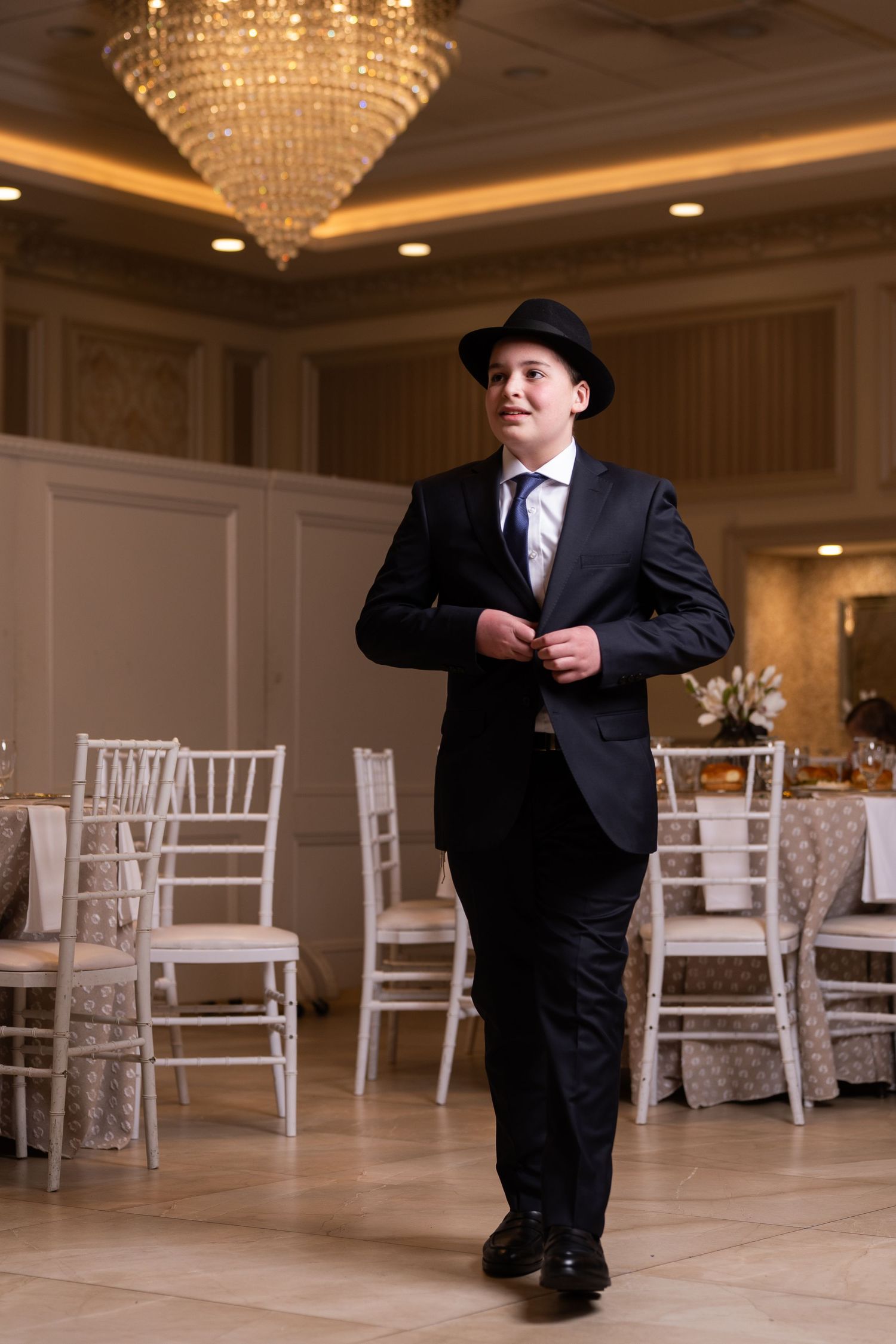 Young boy in black suit and fedora hat buttoning jacket in elegant ballroom with crystal chandelier.
