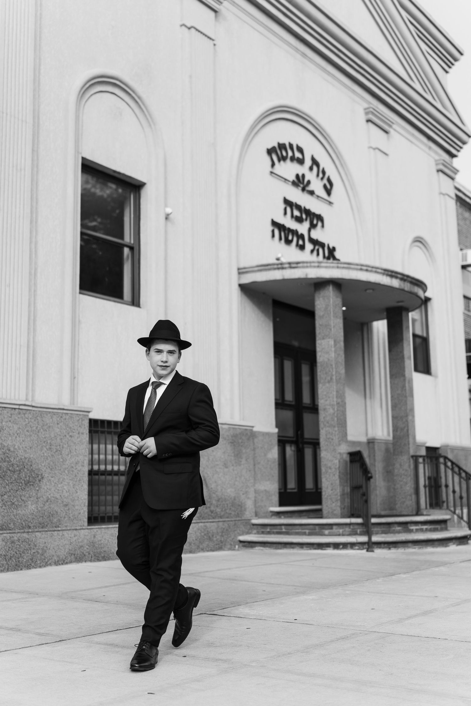 Young man in black suit and fedora stands before a synagogue with Hebrew text in black and white photo.