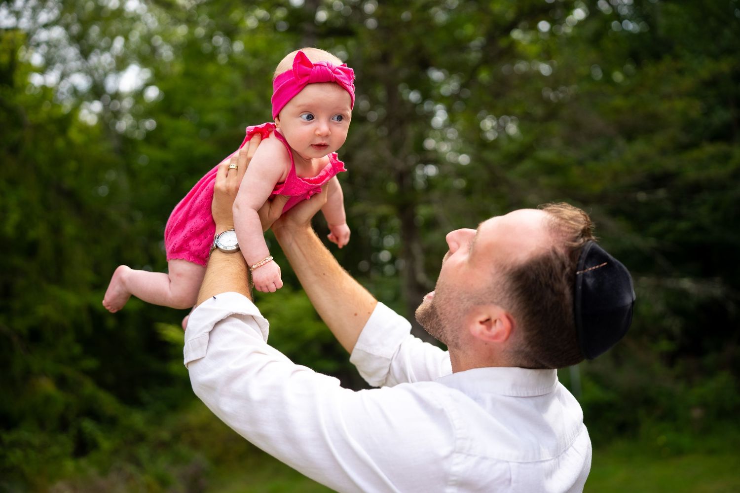 Father joyfully lifting smiling baby girl in pink dress outdoors among green trees.