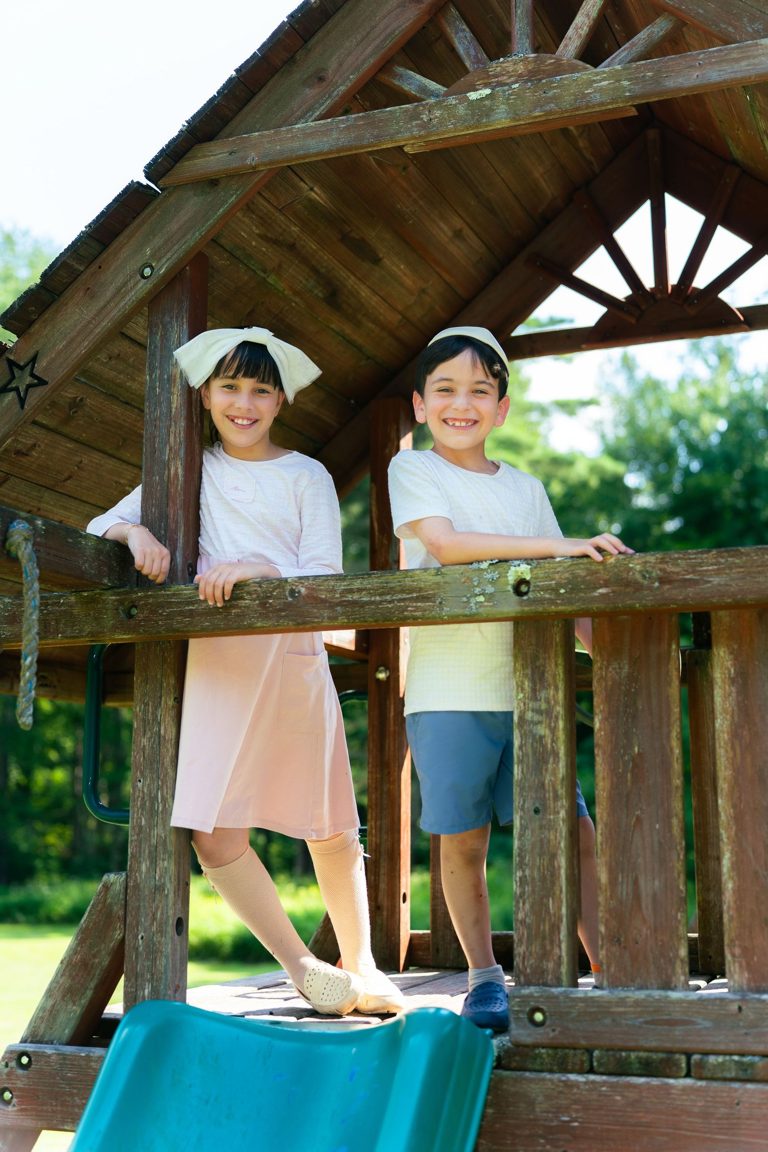 Two smiling children standing on a wooden playground structure on a sunny day.