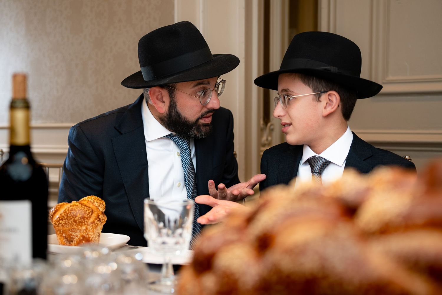 People in black fedoras and suits engaged in conversation at an indoor gathering.