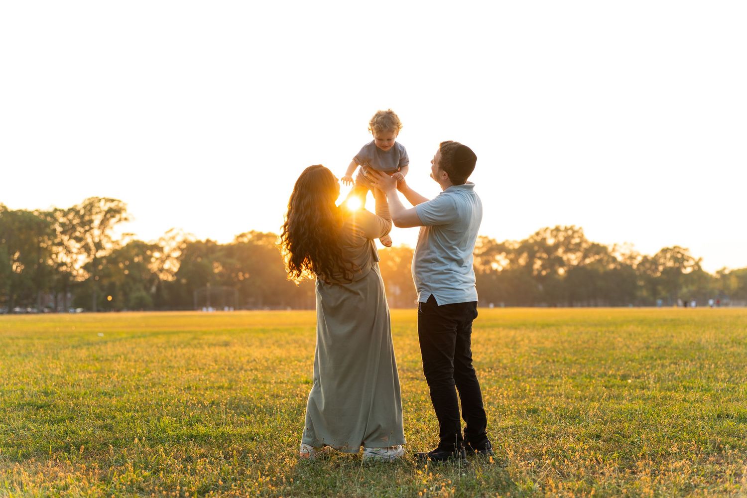 Family silhouettes stand together in a golden field at sunset as sunlight streams through the peaceful scene.