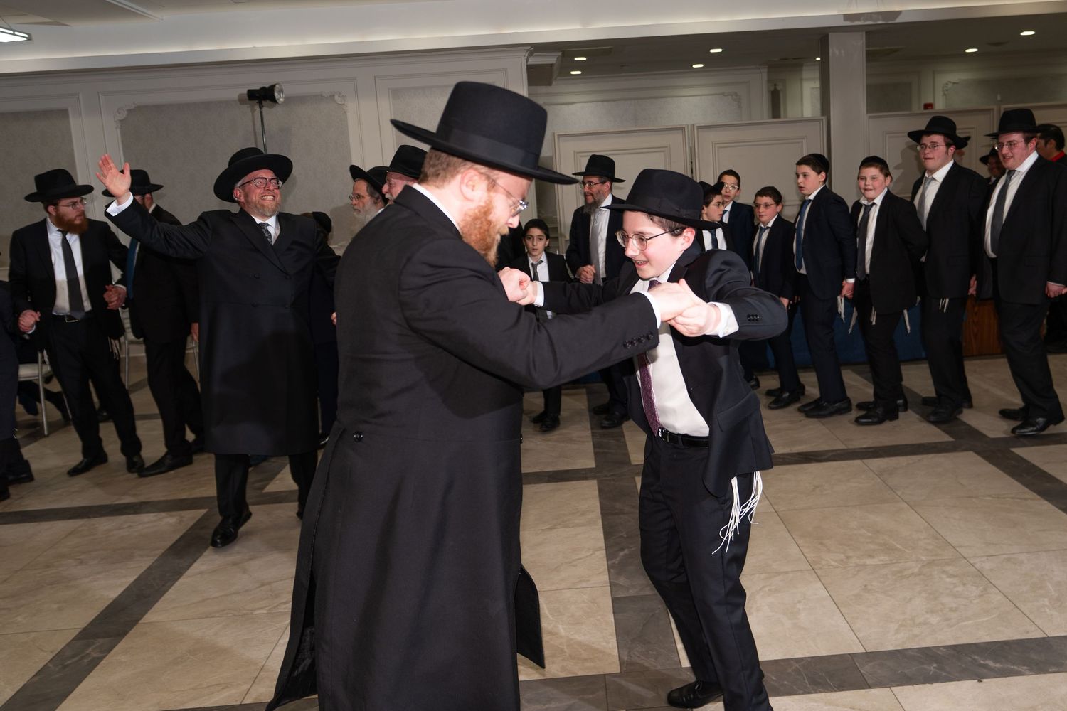 Orthodox Jewish men and boys in black suits and hats dancing together at a celebration.