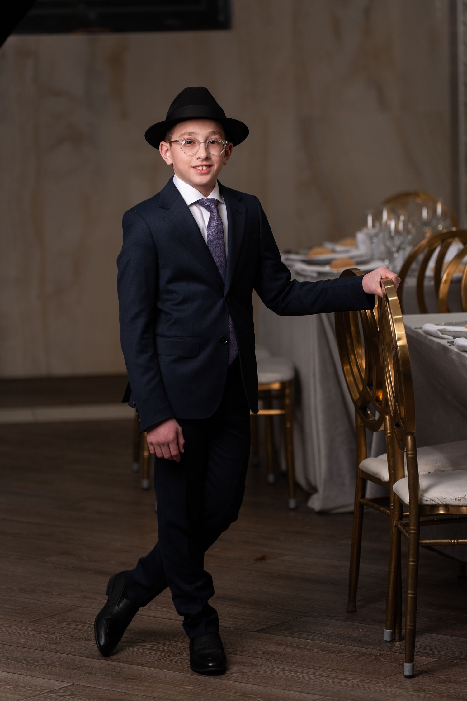 A figure in a black suit and hat poses dramatically in a dining room setting with elegant chairs and tables.