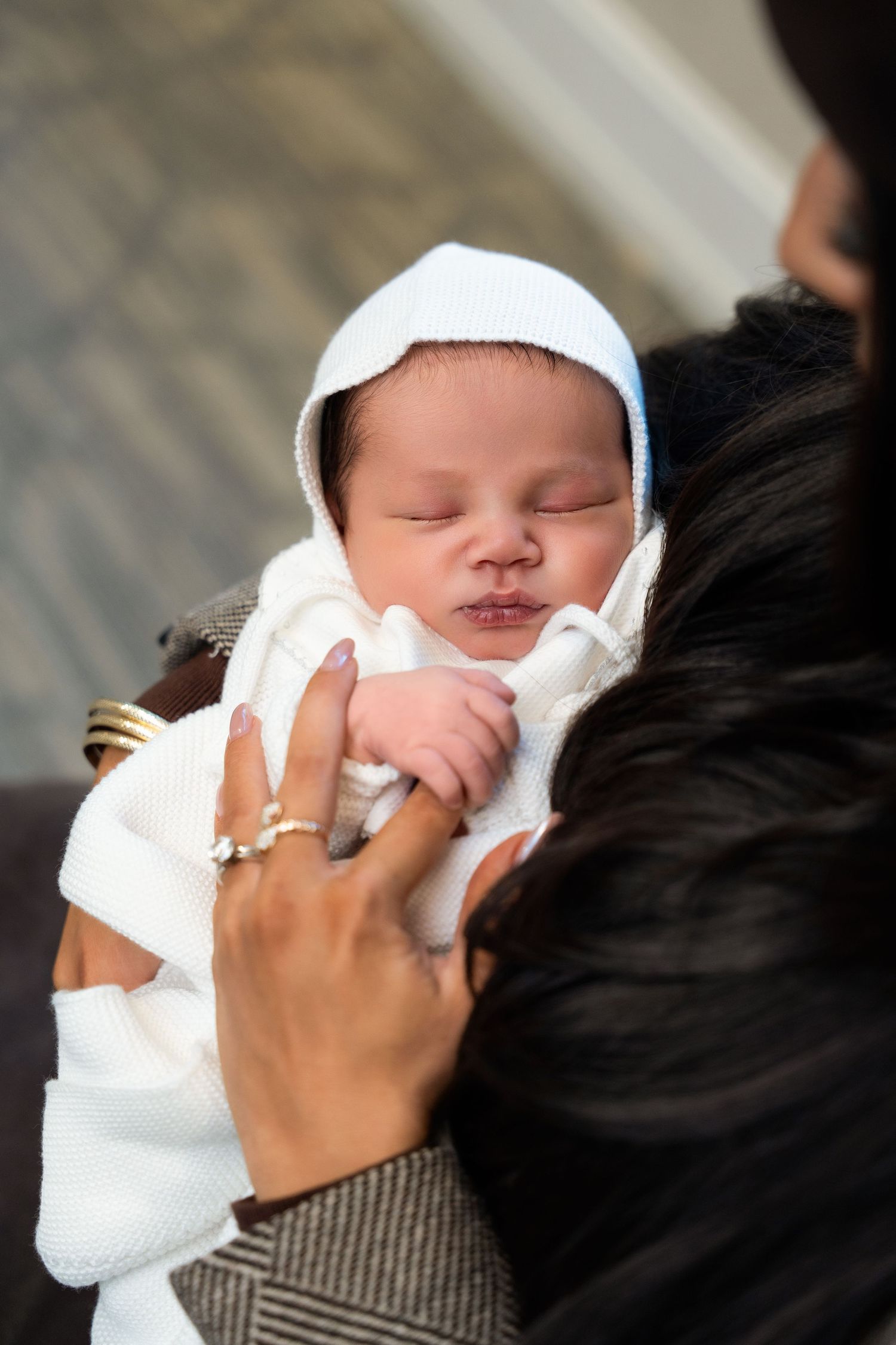 A newborn baby wearing a white cap and blanket is held lovingly while sleeping peacefully.