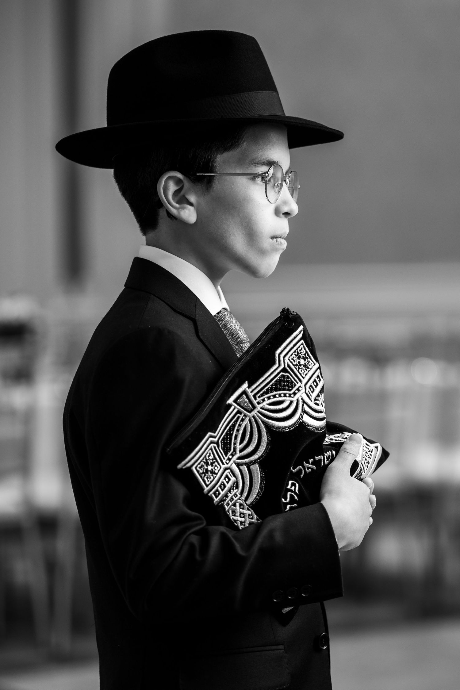 A black and white profile view of a young boy in formal attire and hat holding a patterned bow tie.