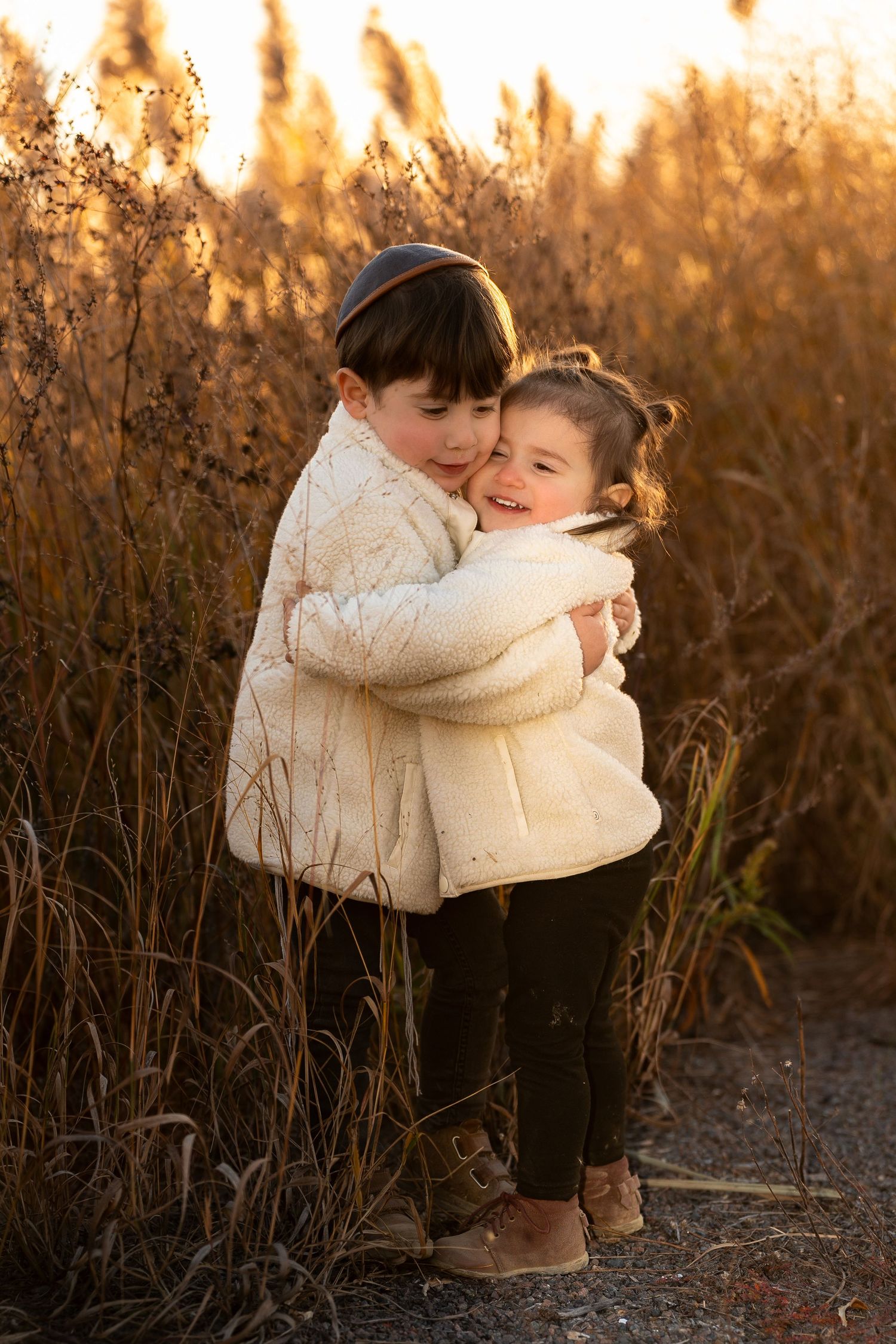 Two children in cozy white sweaters sharing a warm embrace at sunset among tall grass.
