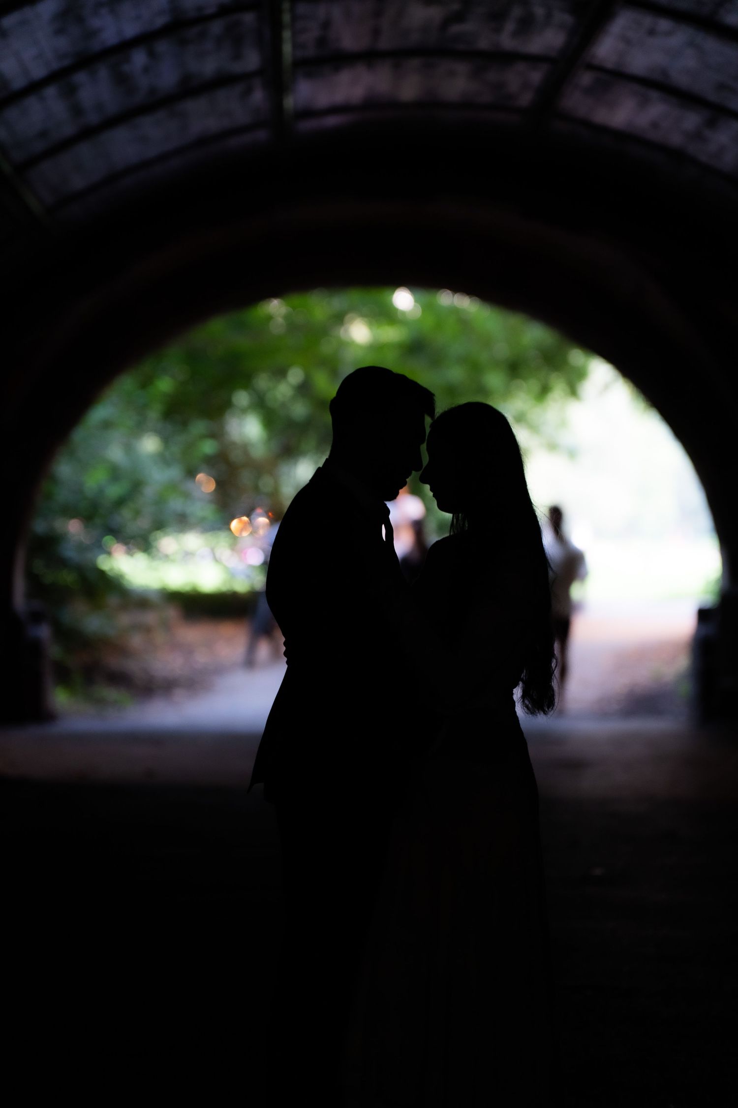 Romantic silhouette of a couple embracing in a stone tunnel arch with lush green trees in the background.