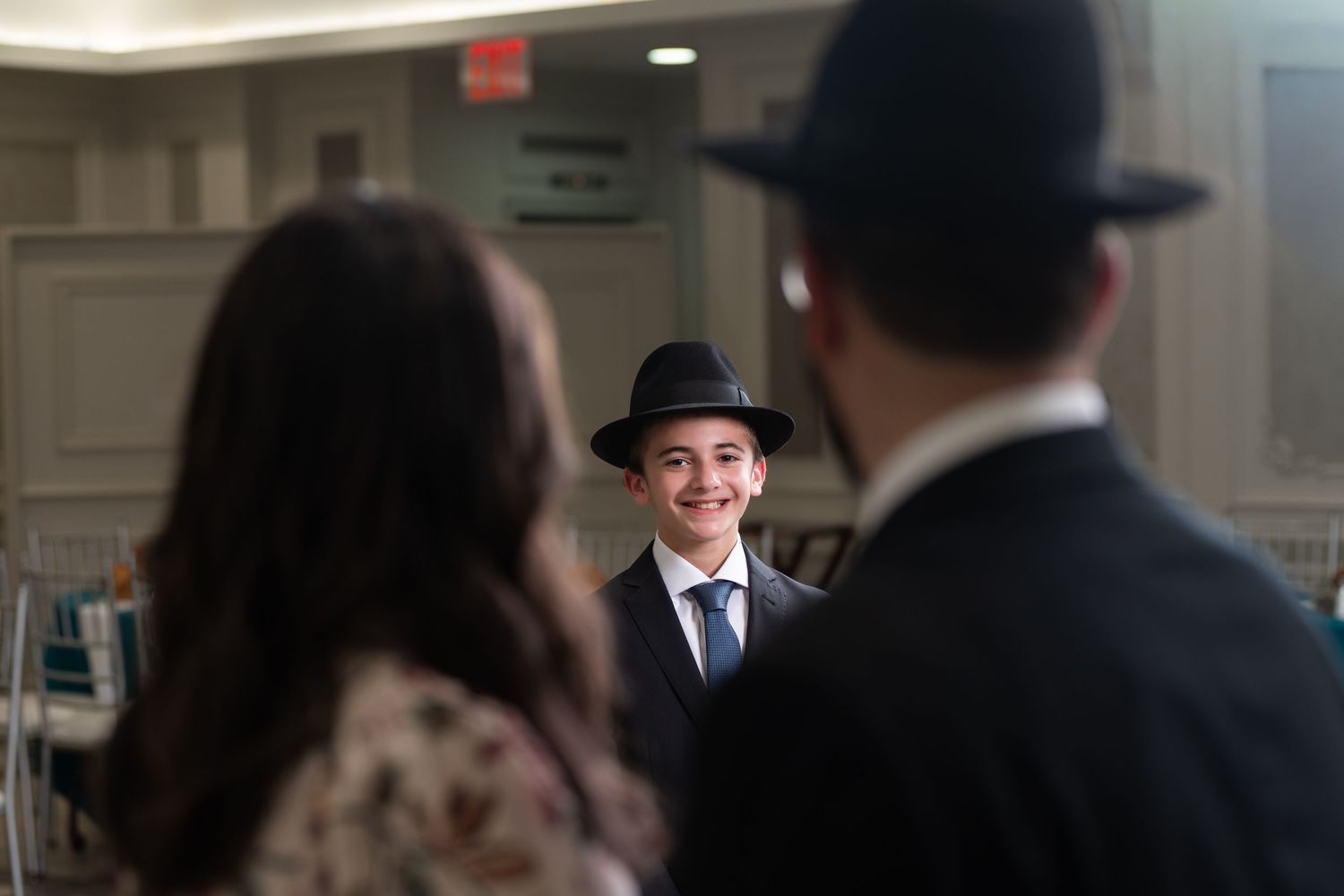 A smiling boy in a black fedora hat and suit at a Bar Mitzvah celebration, greeted by guests in formal attire.