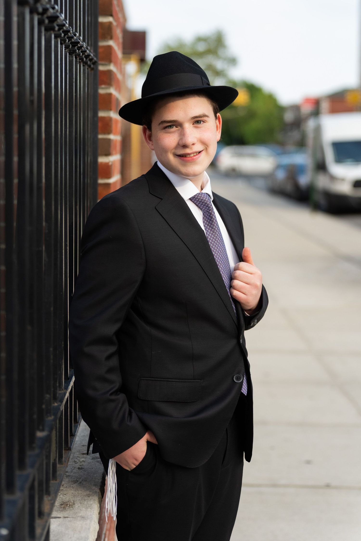 Young man in black suit, tie, and fedora hat smiling on urban sidewalk.