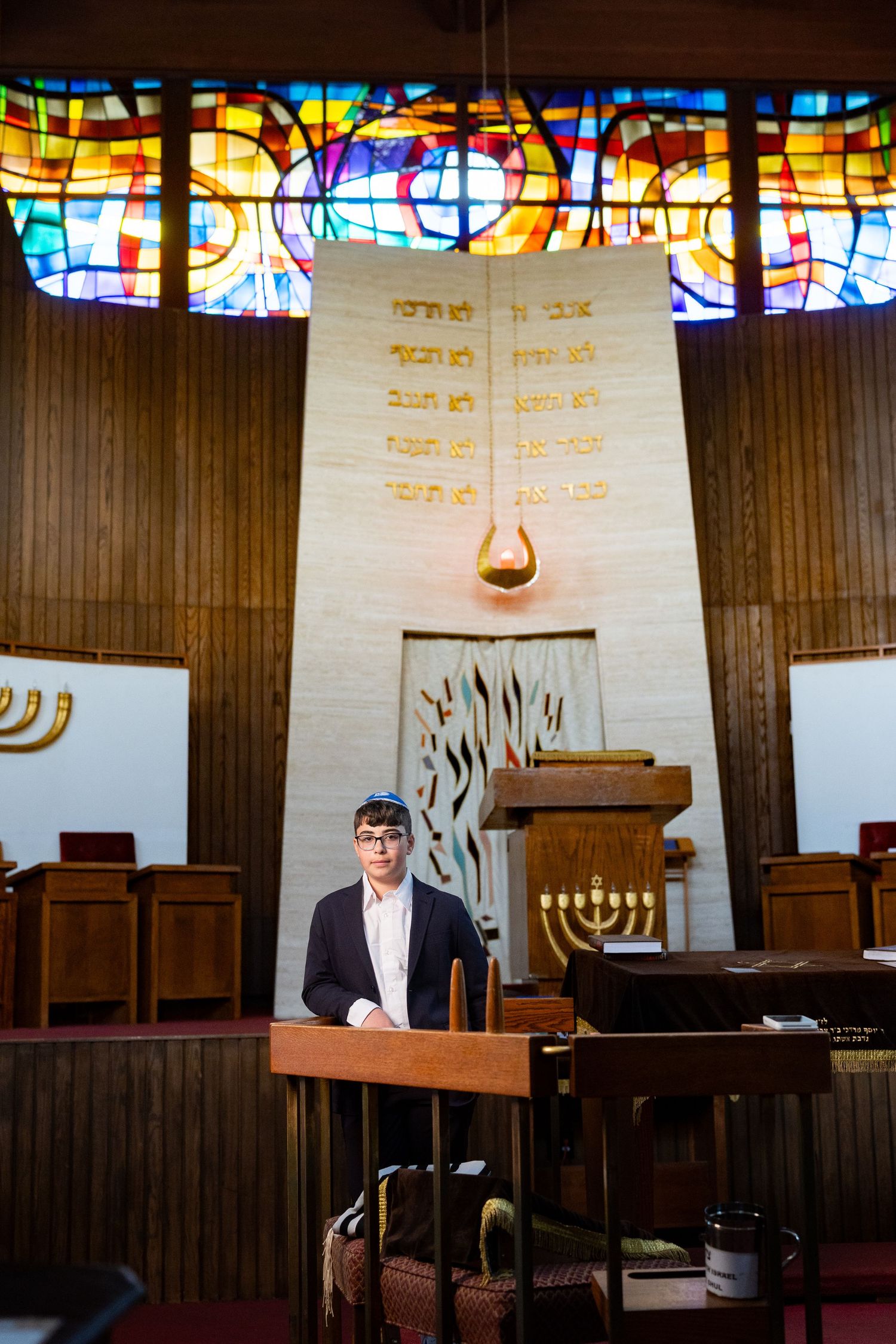 Man standing at podium in synagogue with menorah, Hebrew text backdrop, and colorful stained glass windows.