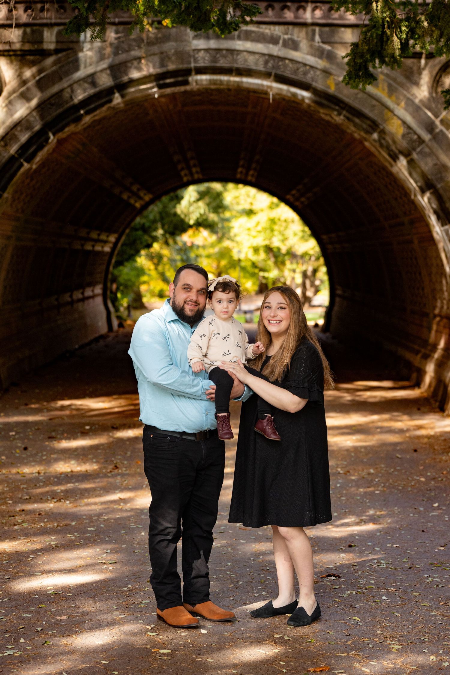 A family of three shares a tender moment under a stone bridge tunnel in a sunlit park setting.