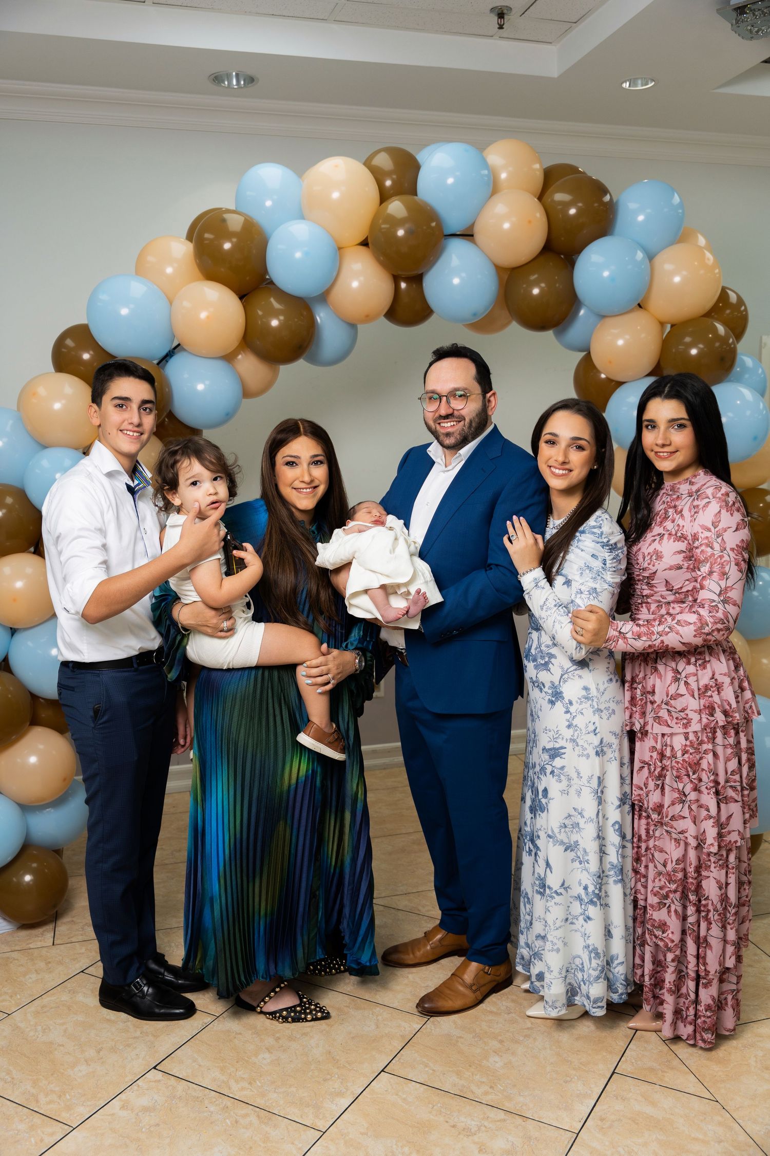 A group of people pose in front of a balloon arch decorated in brown, peach and blue colors at a celebration event.