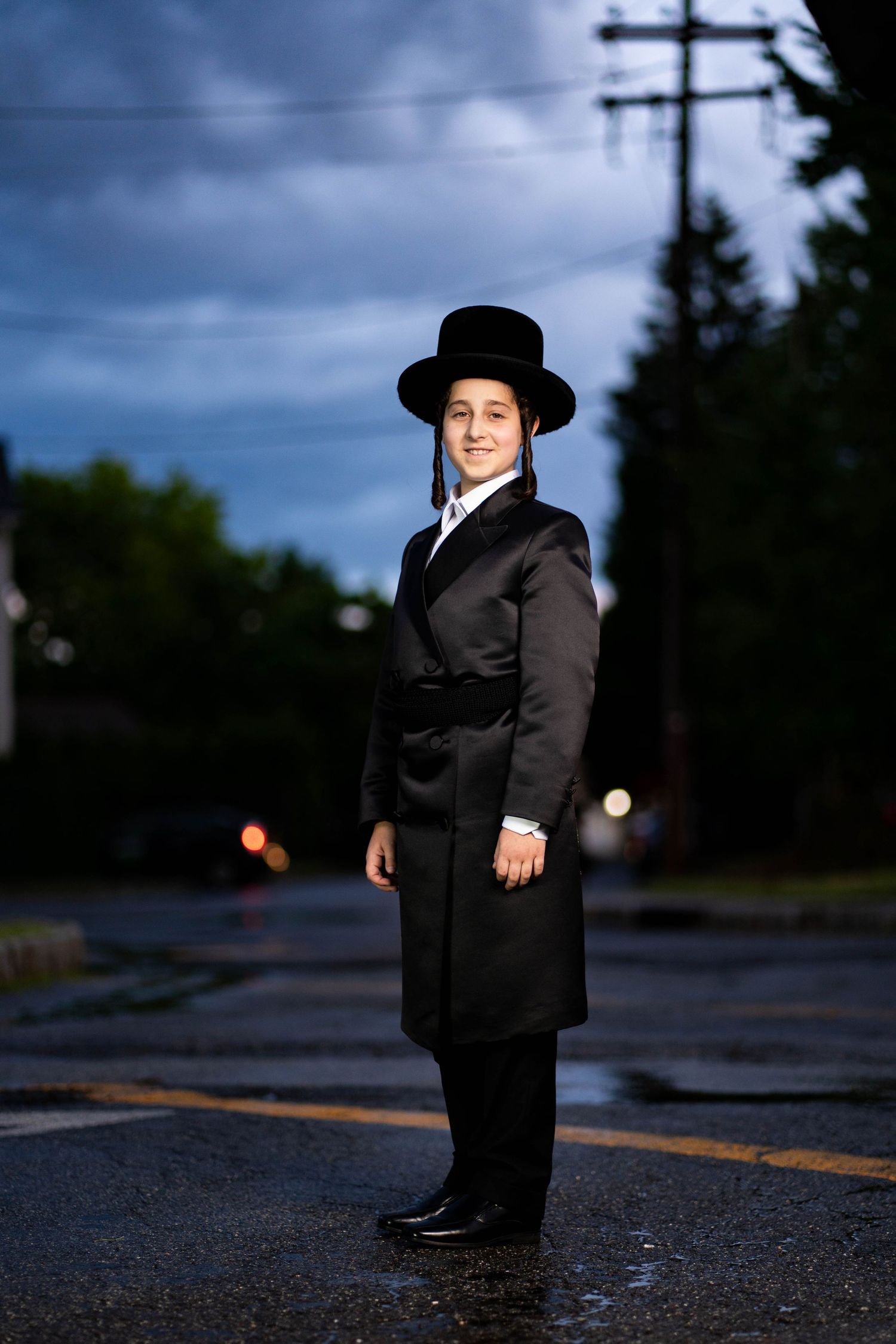 A person in traditional Orthodox Jewish attire stands on a wet street at dusk.