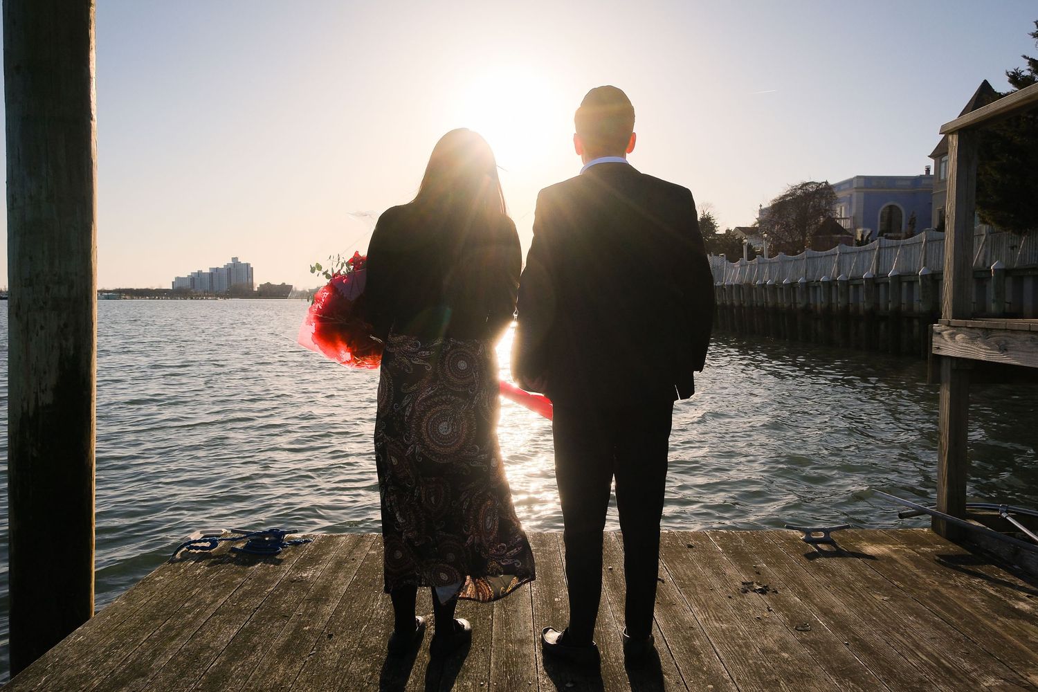 Romantic couple walks along a waterfront pier at sunset with their silhouettes against the golden light.