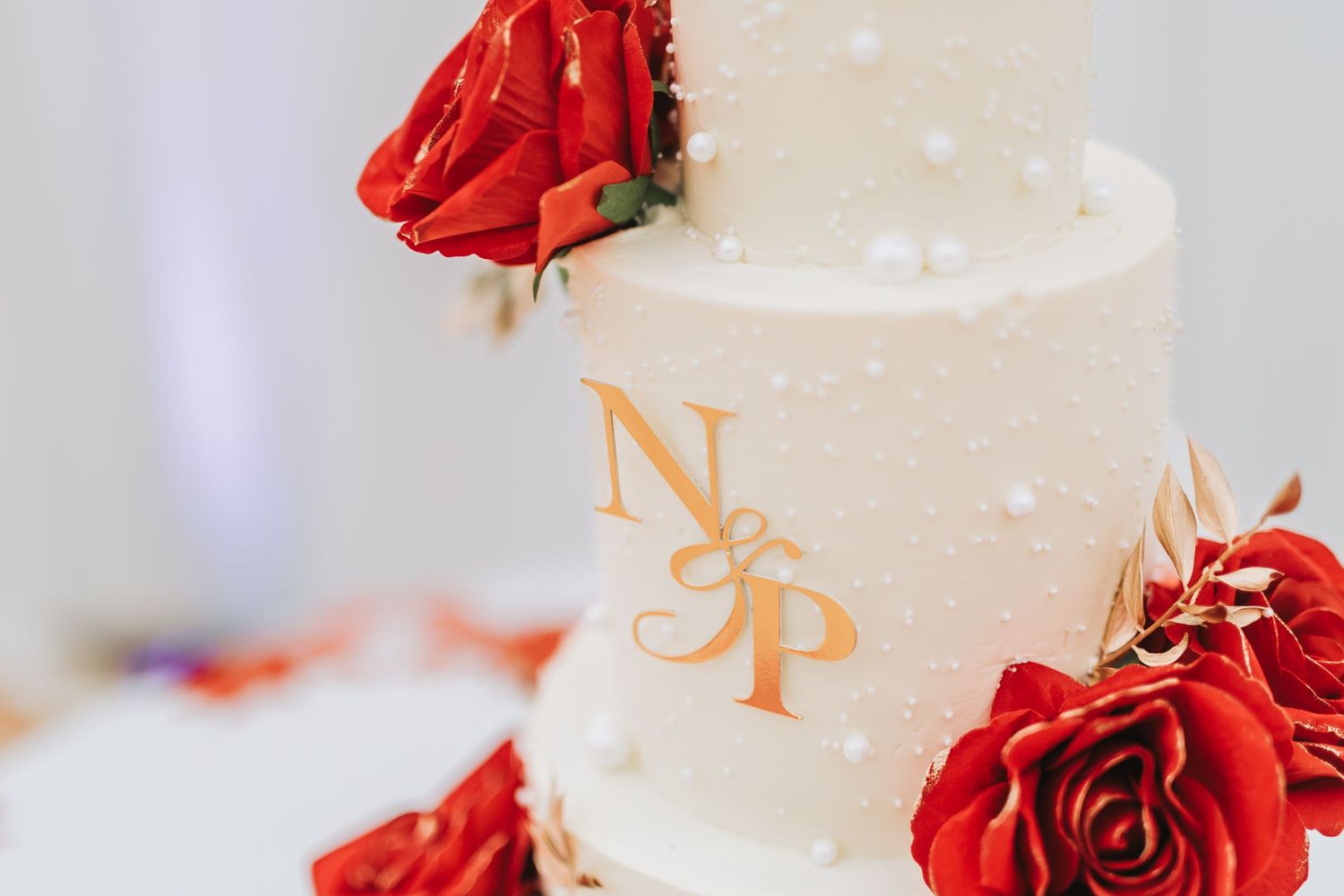 A white wedding cake decorated with vibrant red roses and gold accents against a soft blurred background.