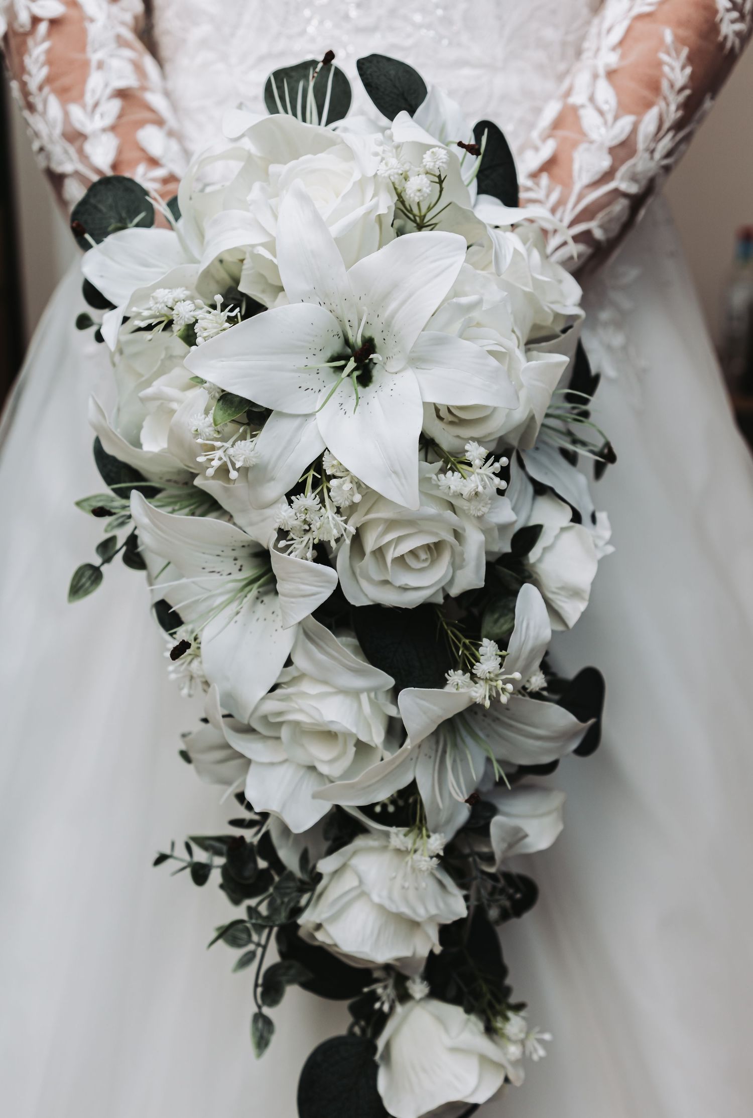 Close-up view of an elegant white wedding bouquet featuring lilies, roses, and green foliage against a white dress background.