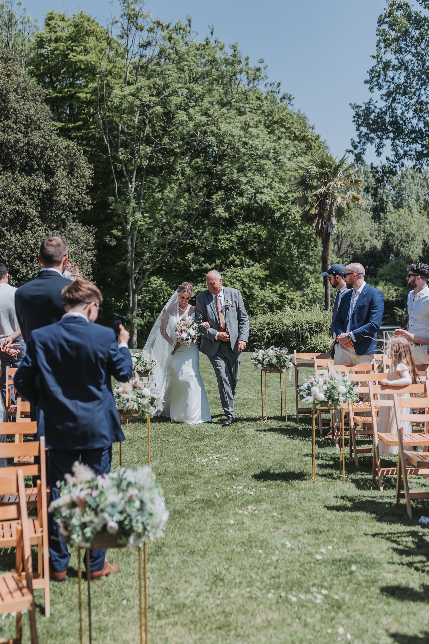 Outdoor garden wedding ceremony with wooden chairs and floral arrangements lining the aisle on a sunny summer day.