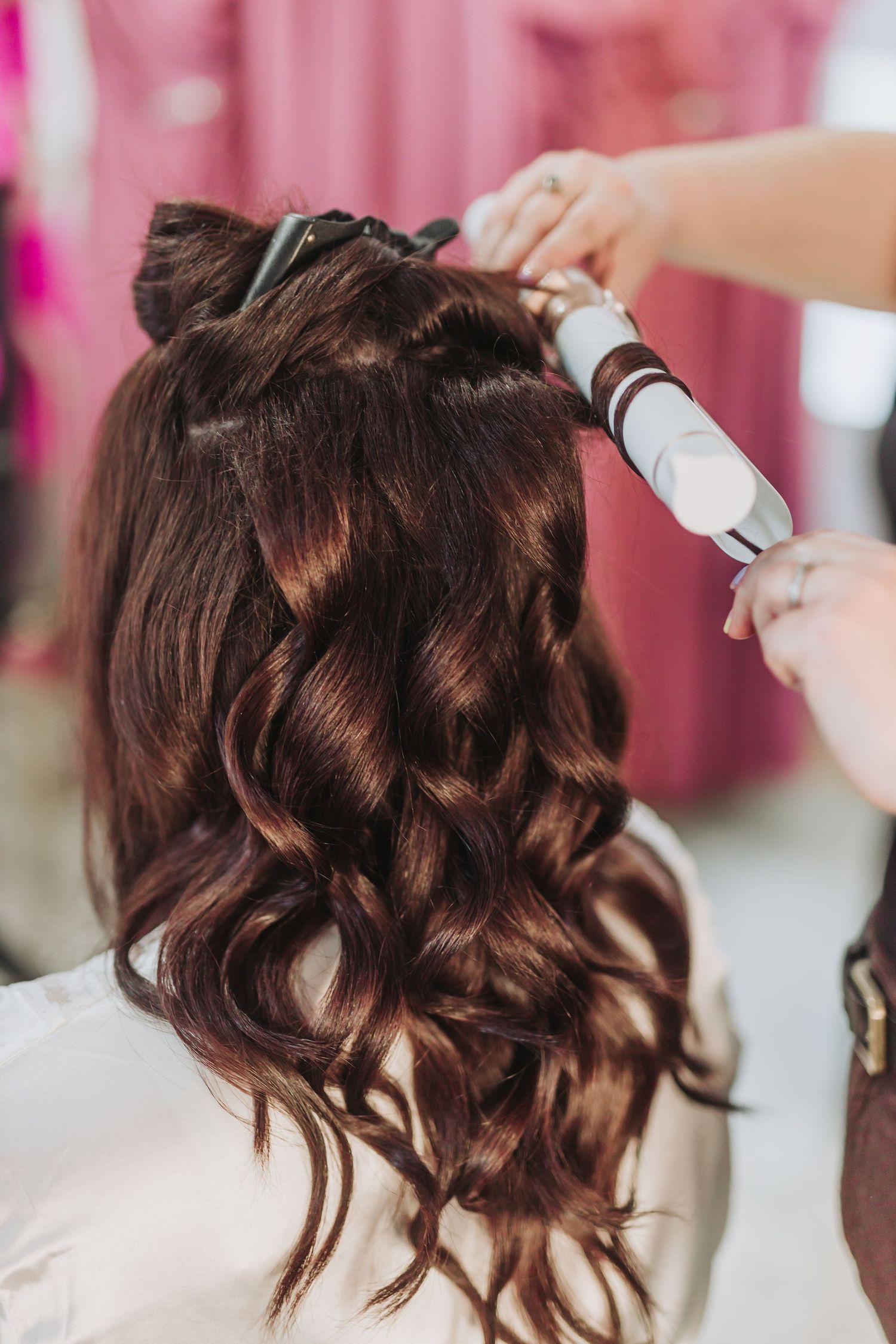 Hair stylist spraying hairspray on dark brown curled hair while styling against pink background.