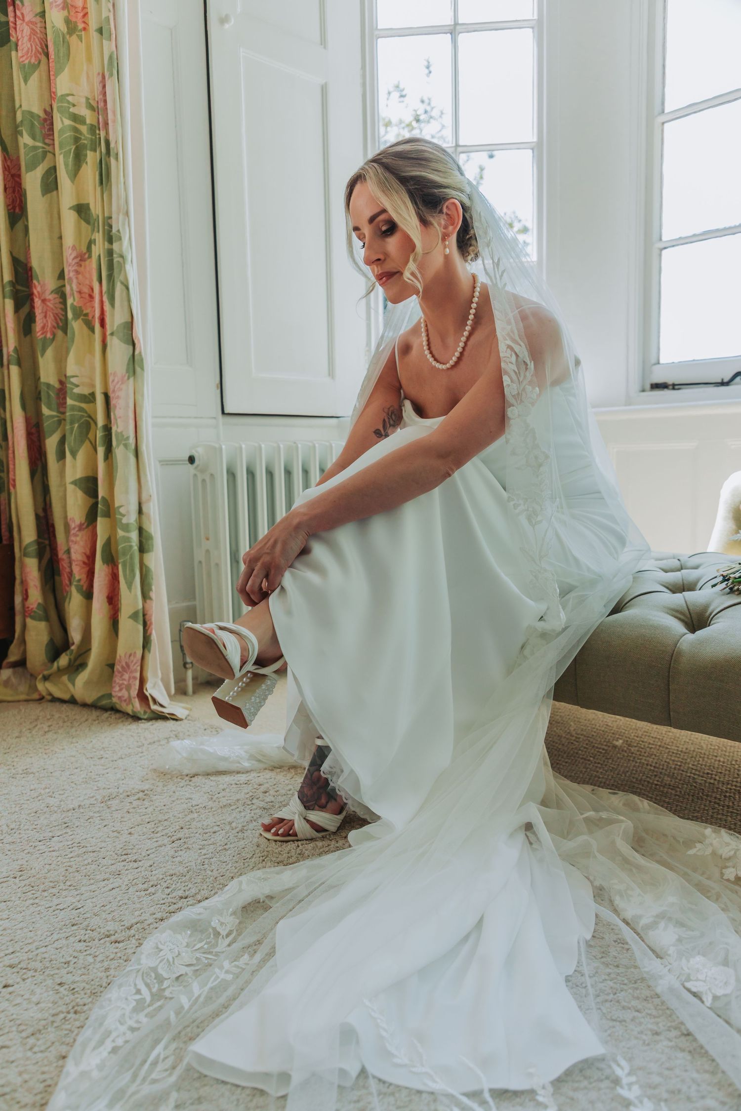 A bride in a flowing white wedding dress adjusts her shoes while standing in a sunlit room with floral curtains.