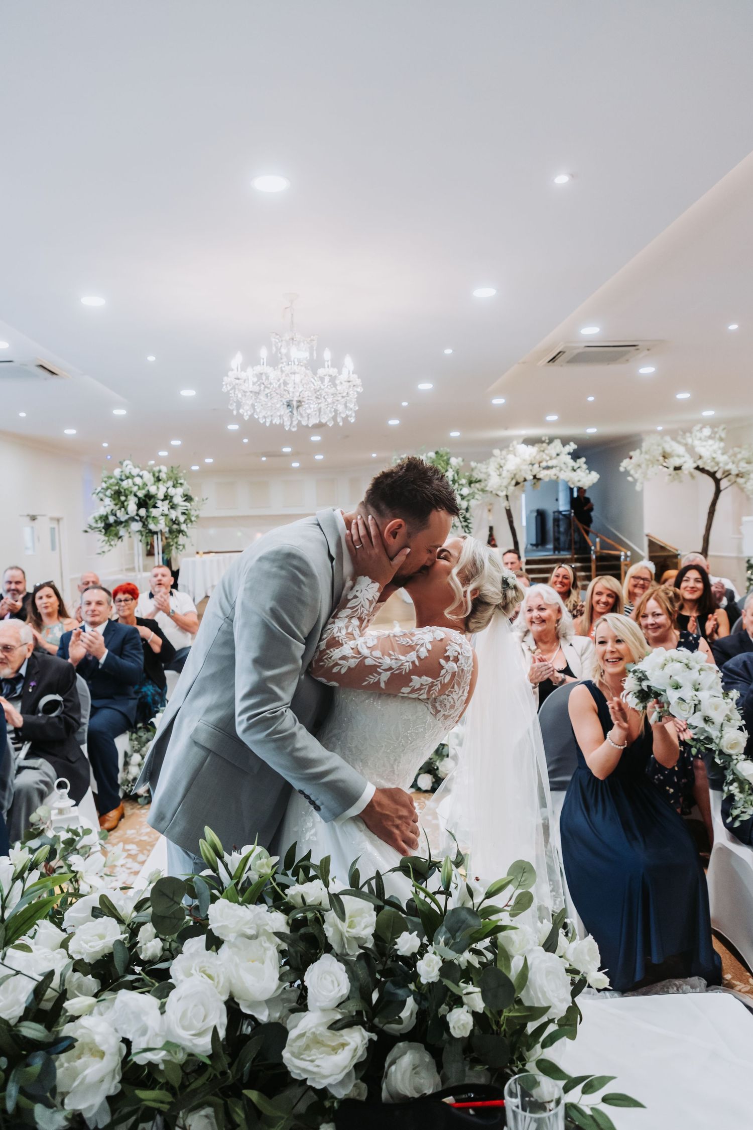 Elegant wedding reception scene with white floral arrangements and guests celebrating in a bright ballroom.