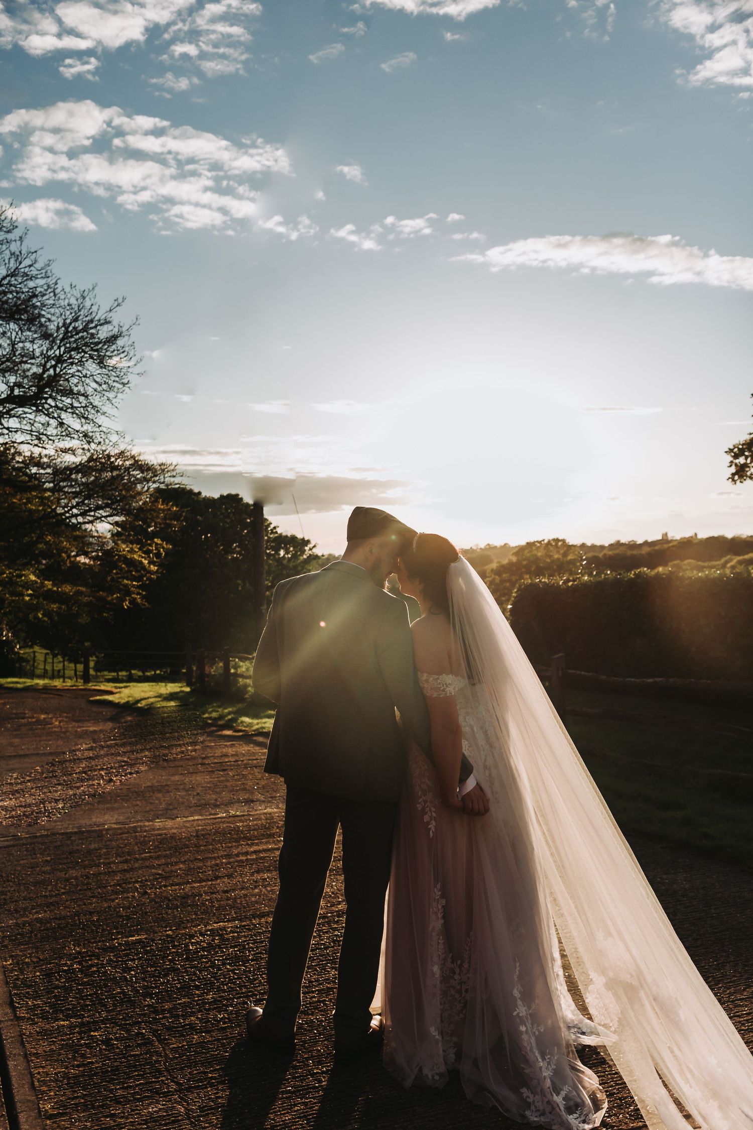 A bride and groom share an intimate moment at sunset on a dirt path surrounded by trees and dramatic sky.
