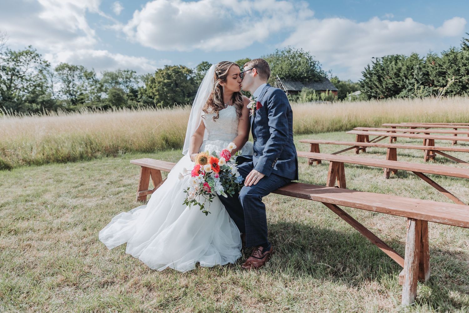 Newlyweds embrace on wooden benches surrounded by tall grass and blue skies on their wedding day.