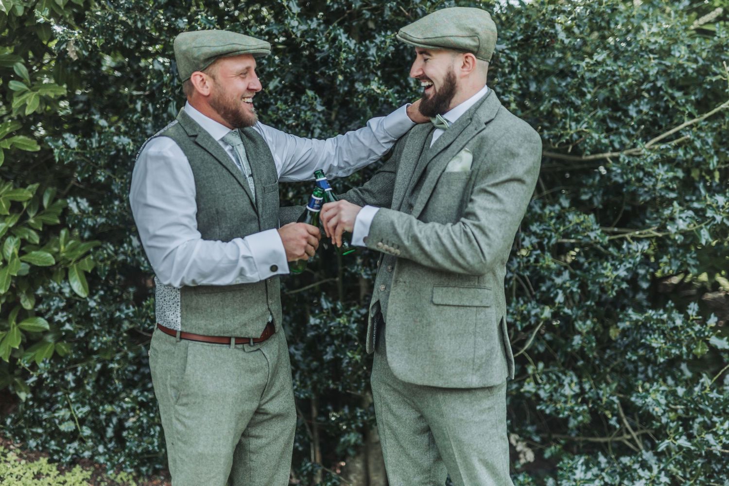 Two people in matching sage green suits share a celebratory moment in a garden setting.